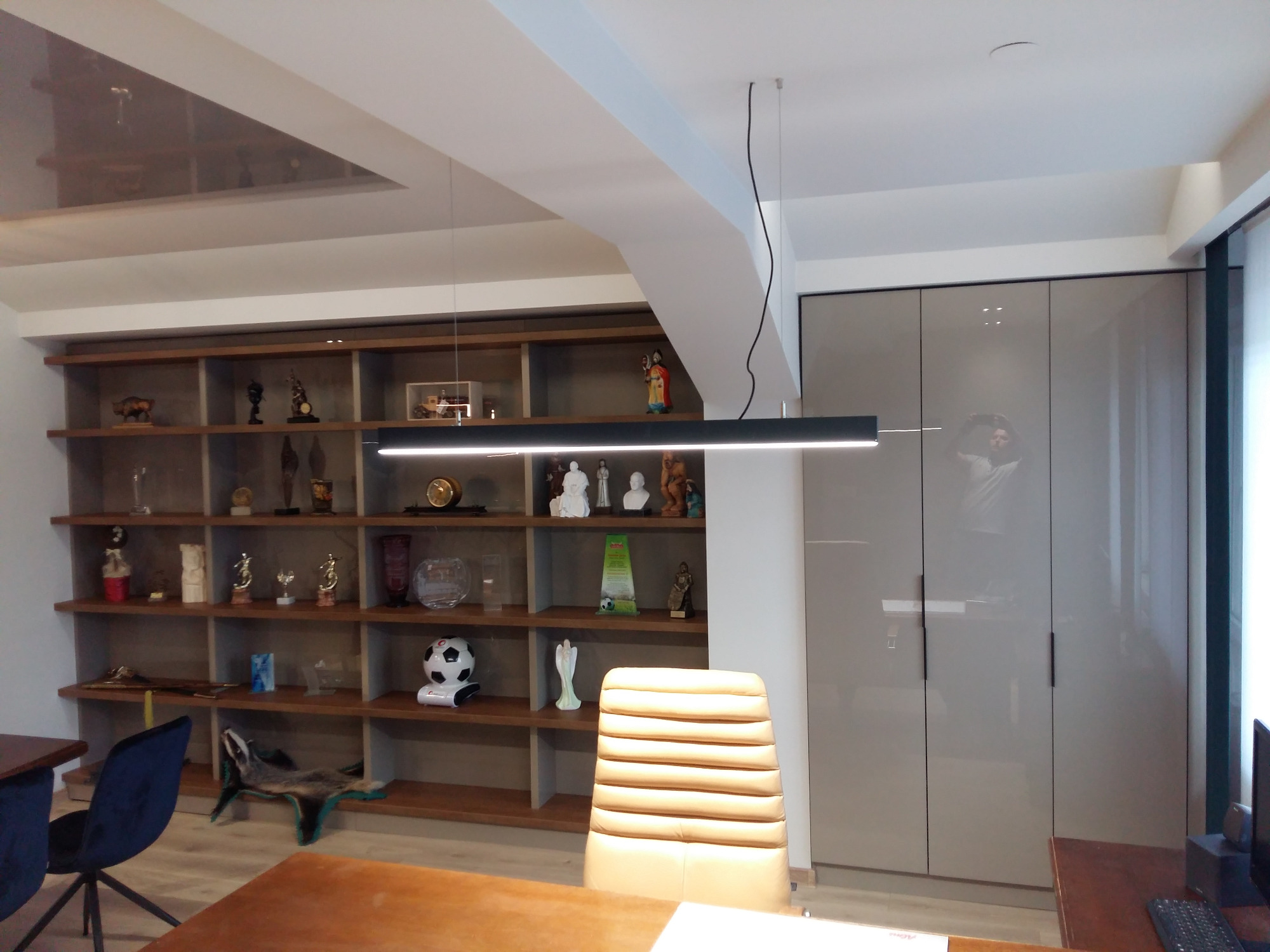 Nowoczesne biuro z regałem na dekoracje i beżową szafą z połyskiem, oświetlone podłużną lampą LED.