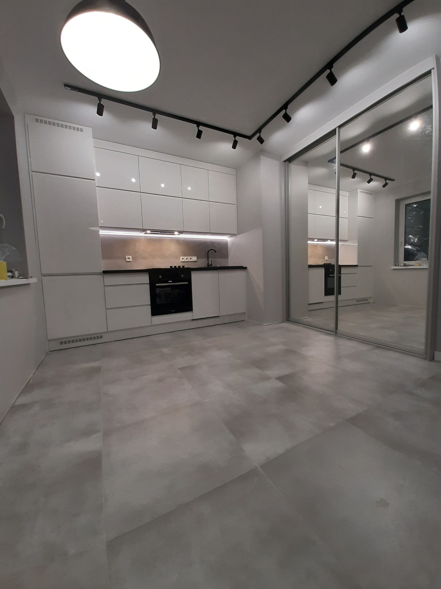 Nowoczesna, minimalistyczna kuchnia z białymi, lakierowanymi frontami szafek, czarnym blatem i oświetleniem punktowym na szynoprzewodzie. Obok kuchni szafa wnękowa z lustrzanymi drzwiami przesuwnymi.