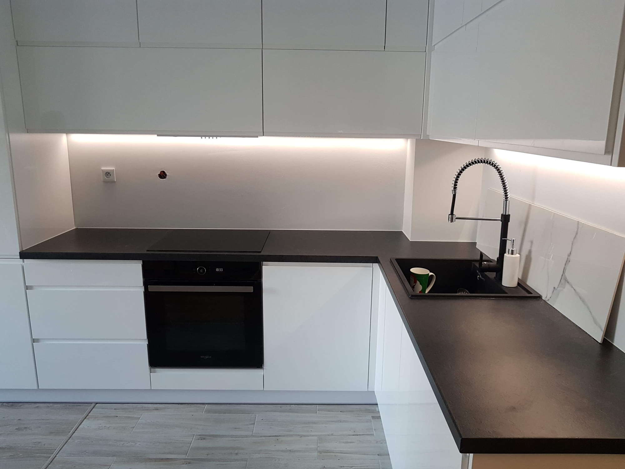 Nowoczesna biała kuchnia z czarnym blatem, oświetleniem LED podszafkowym i czarną armaturą. Widoczny fragment płytek imitujących marmur.