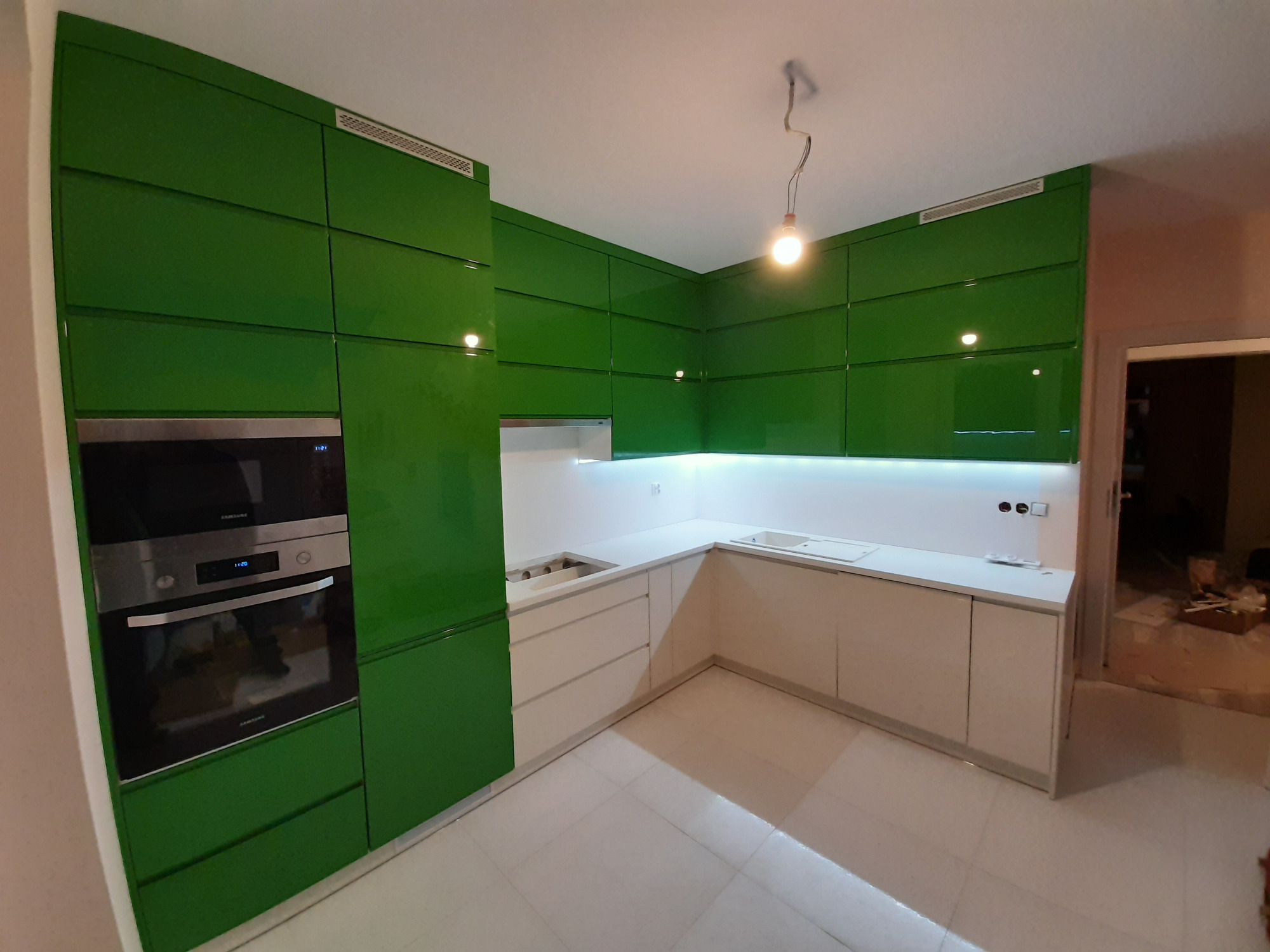 Nowoczesna kuchnia z błyszczącymi, zielonymi szafkami i białymi blatami, oświetlona lampą wiszącą, z wbudowanym piekarnikiem Samsung.