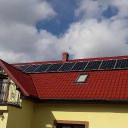 Czerwony dach domu jednorodzinnego z zamontowanymi panelami słonecznymi, widoczne okno dachowe i balkon z ozdobną balustradą, na tle błękitnego nieba z chmurami.