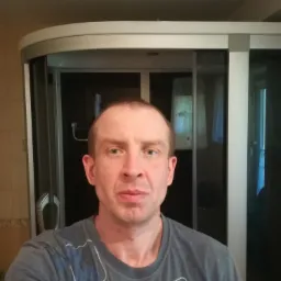 Mężczyzna w szarej koszulce na tle łazienki z kabiną prysznicową z przesuwanymi drzwiami.