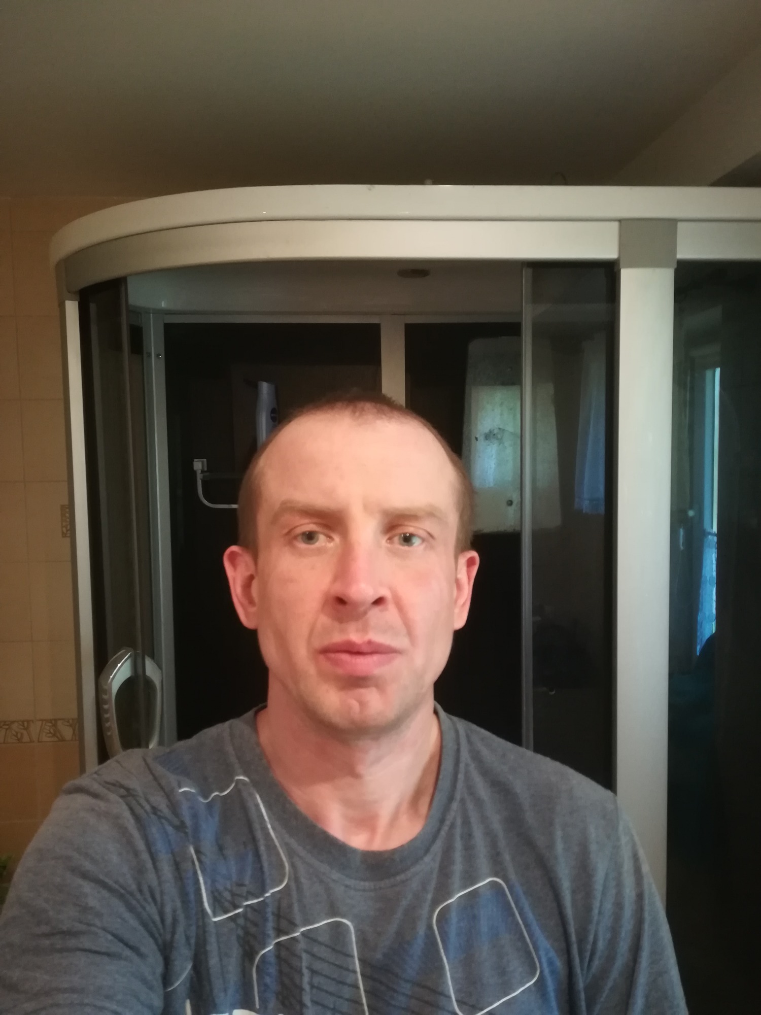 Mężczyzna w szarej koszulce na tle łazienki z kabiną prysznicową z przesuwanymi drzwiami.