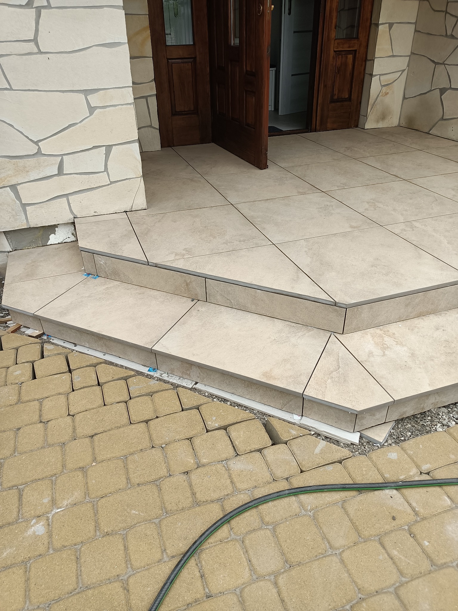 Wyłożone jasnymi płytkami schody wejściowe do domu z otwartymi drewnianymi drzwiami, widoczne niedokończone obrzeże z kostki brukowej i fragment węża ogrodowego.