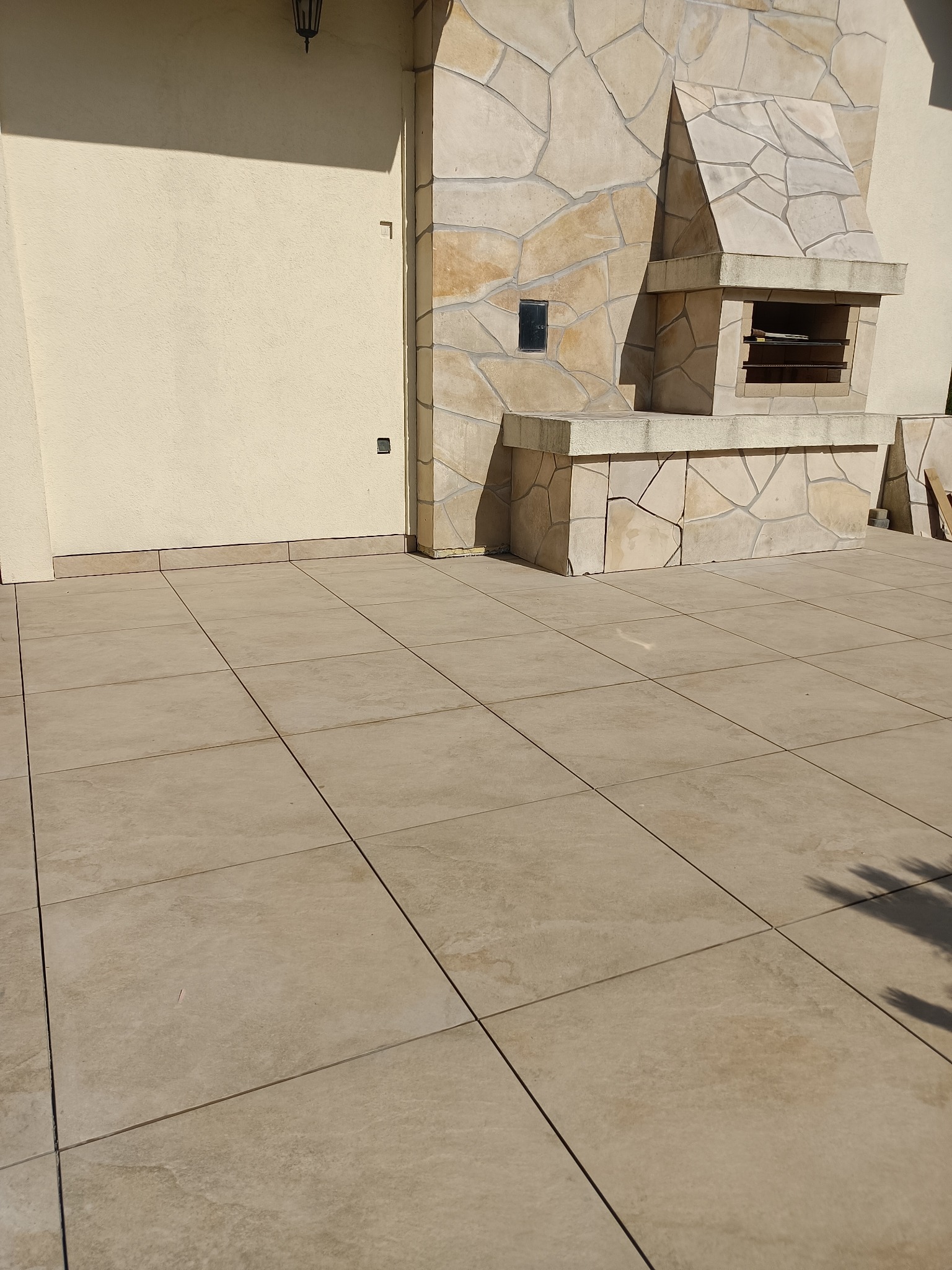 Ułożona jasna, kwadratowa terakota na zewnątrz budynku z kamiennym grillem w tle, widoczne drobne niedoskonałości na powierzchni płytek.