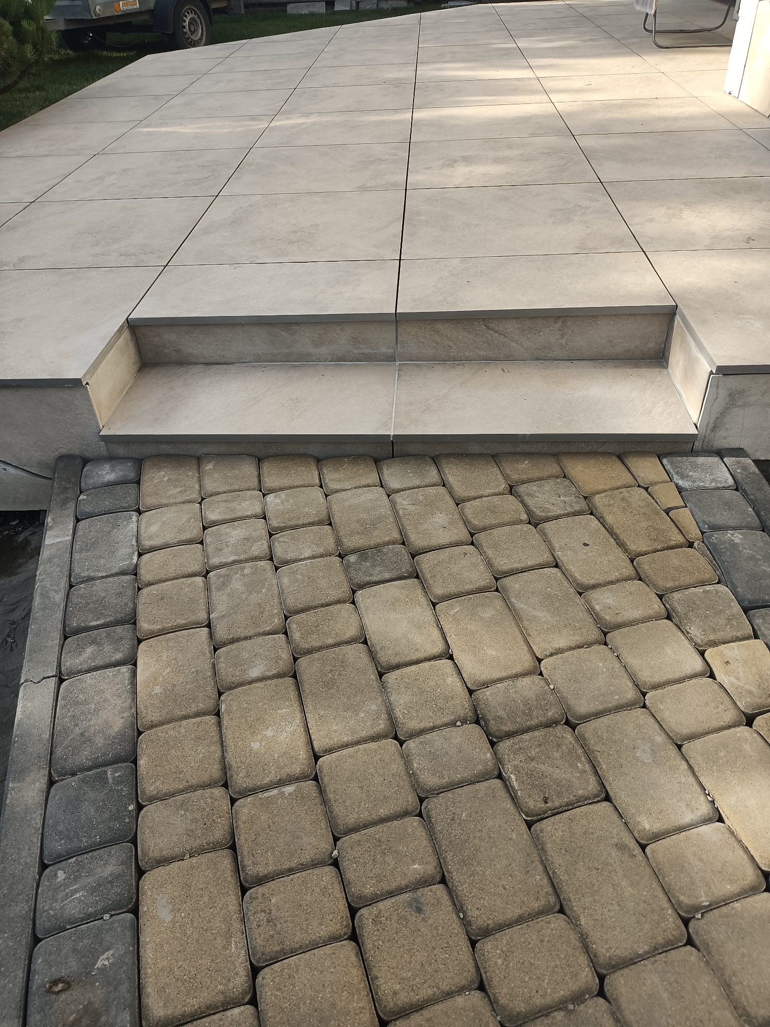 Ułożona kostka brukowa przechodząca w stopień i taras wyłożony płytkami w odcieniach beżu i szarości.