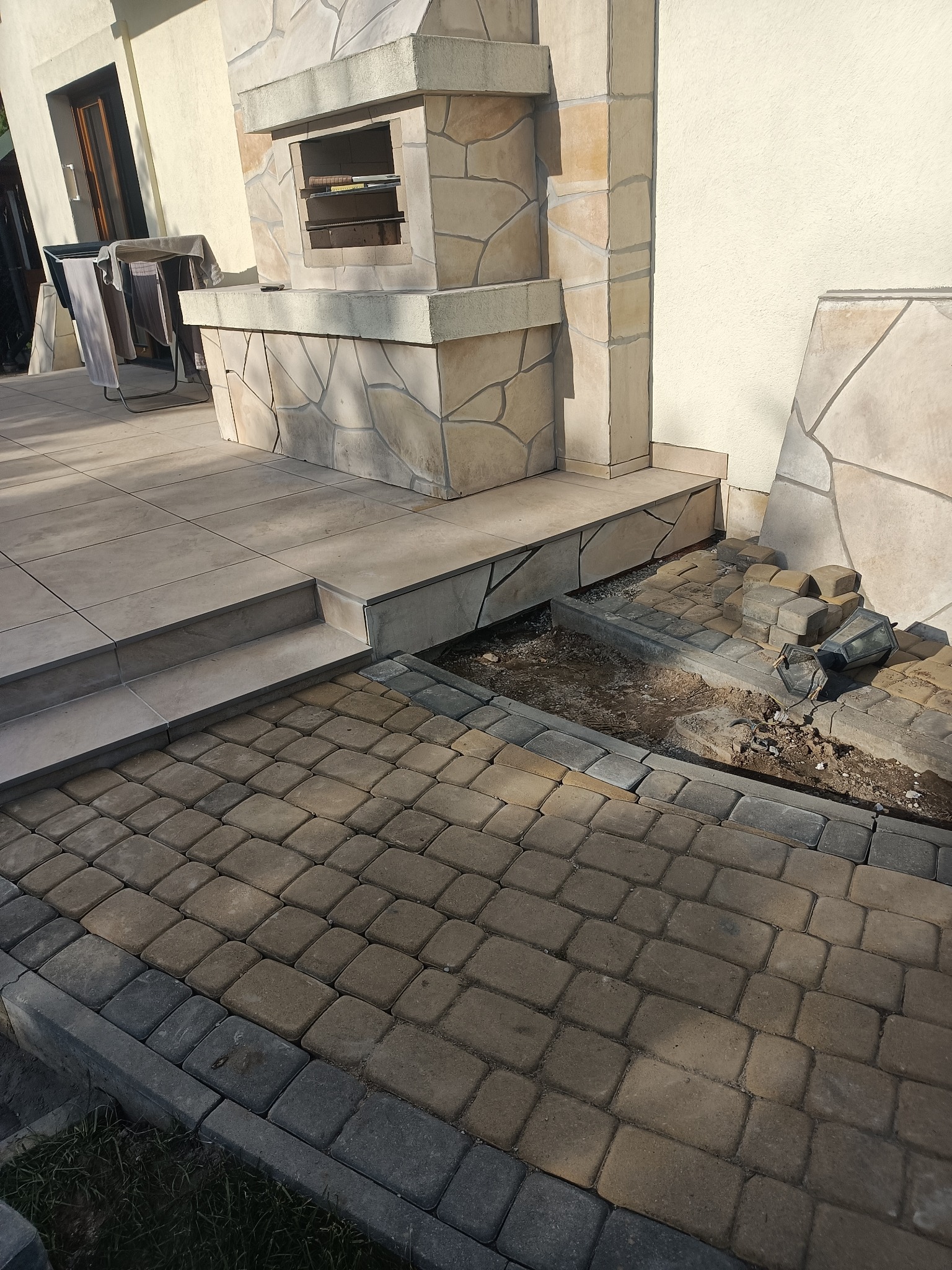 Układanie kostki brukowej na posesji w Lublinie, widoczny fragment nowej nawierzchni oraz przygotowane podłoże z kostką do ułożenia, tło stanowi fragment elewacji budynku z kamiennym grillem.