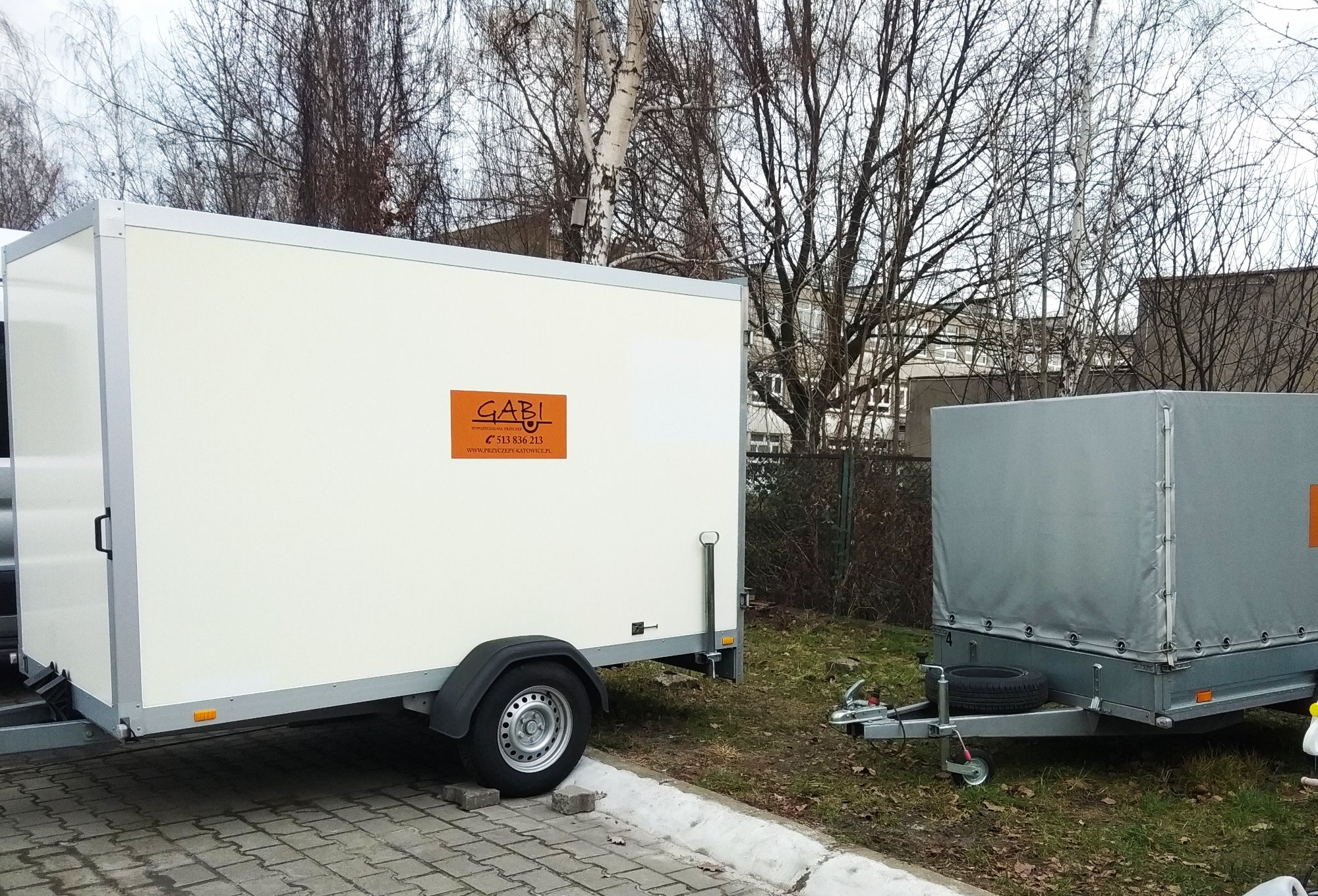 Dwie przyczepy: biała furgonetka z pomarańczowym logo 'GABI' z numerem telefonu i adresem strony internetowej firmy z Katowic, oraz szara przyczepa z plandeką, stojące na tle drzew i budynku.
