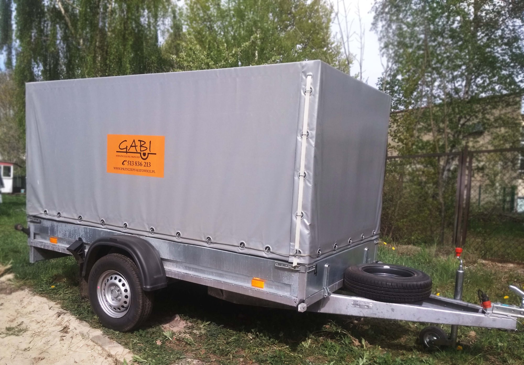 Szara przyczepa towarowa z plandeką, na której widnieje pomarańczowa naklejka z logo firmy GABI i numerem telefonu, zaparkowana na trawie w otoczeniu drzew.
