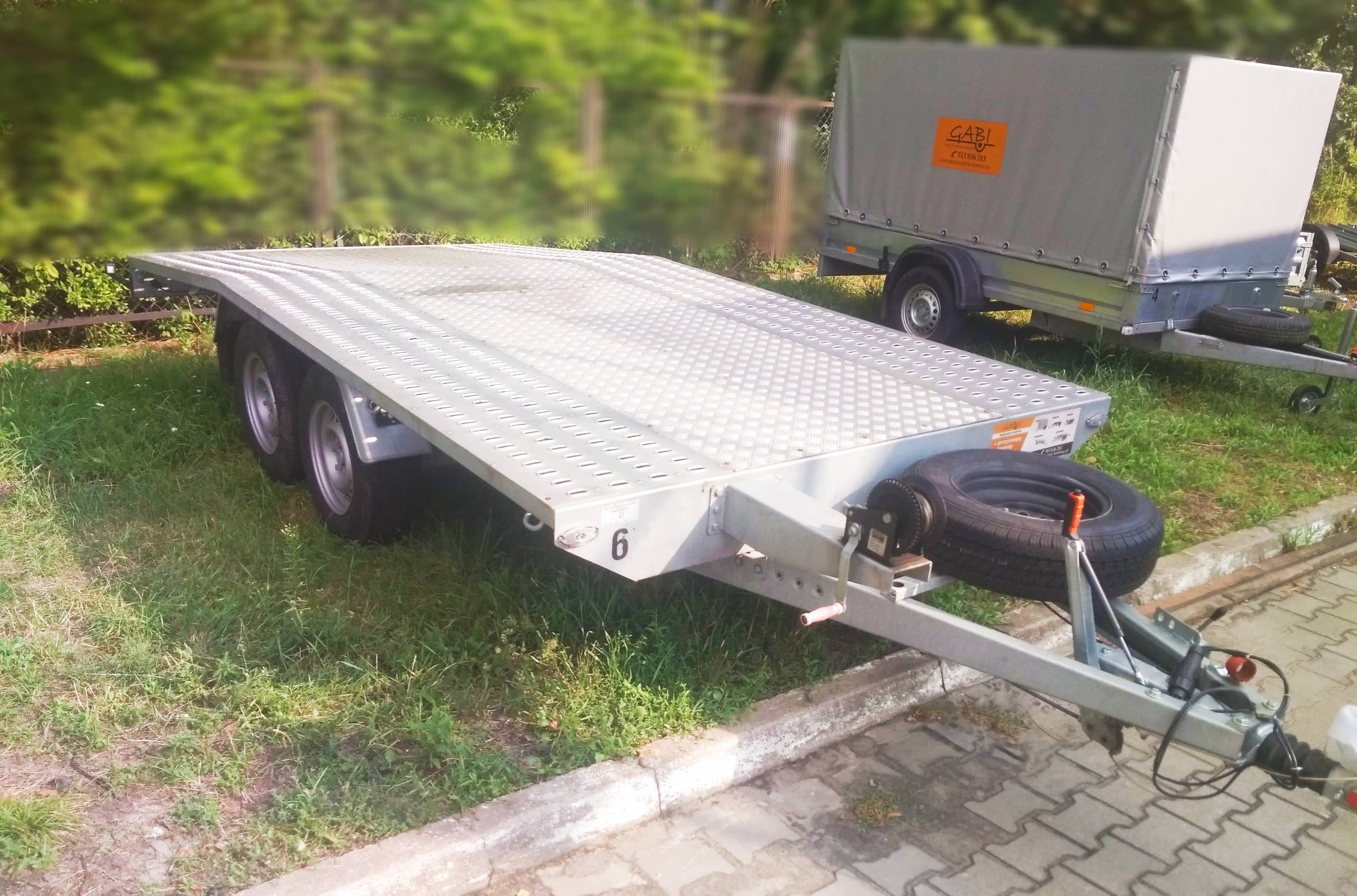 Aluminiowa, dwuosiowa laweta samochodowa z zapasowym kołem, stojąca na trawie, z widoczną wyciągarką ręczną i numerem '6' na boku.
