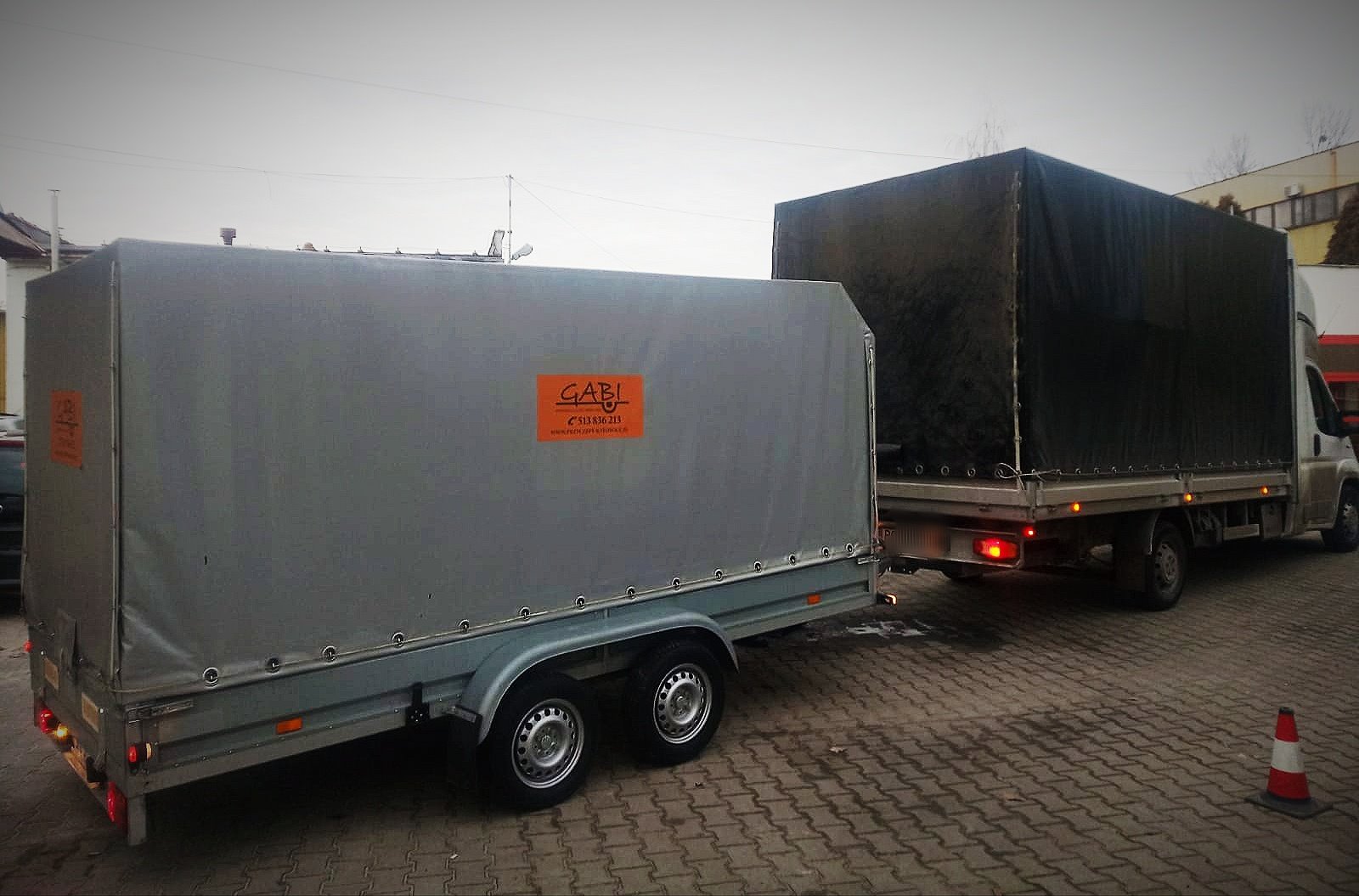 Ciężarówka z czarną plandeką i przyczepa z szarą plandeką na kostce brukowej, pomarańczowa naklejka z logo 'GABI' na przyczepie, stożek ostrzegawczy z boku.