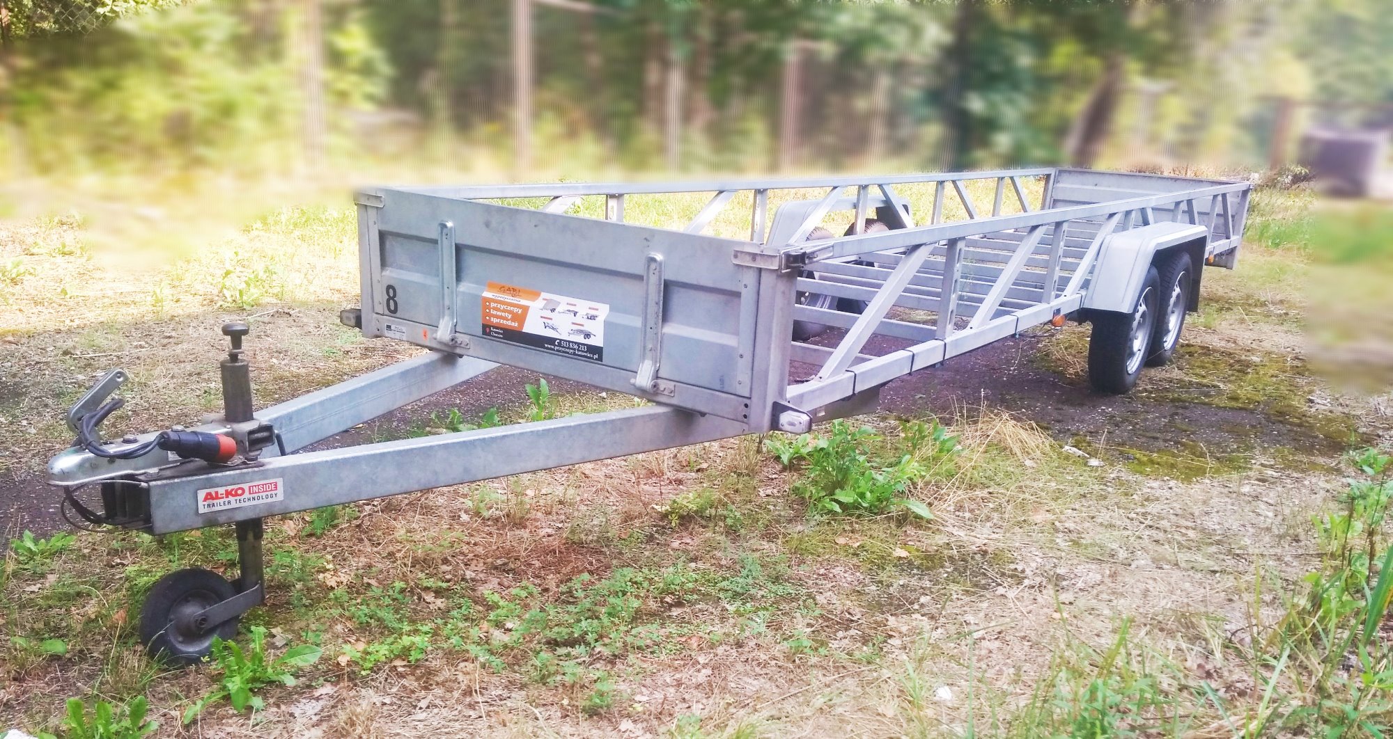 Szara, pusta przyczepa samochodowa z konstrukcją kratownicową na tle zamazanego lasu, widoczny zaczep z kołem podporowym i naklejki informacyjne na boku.