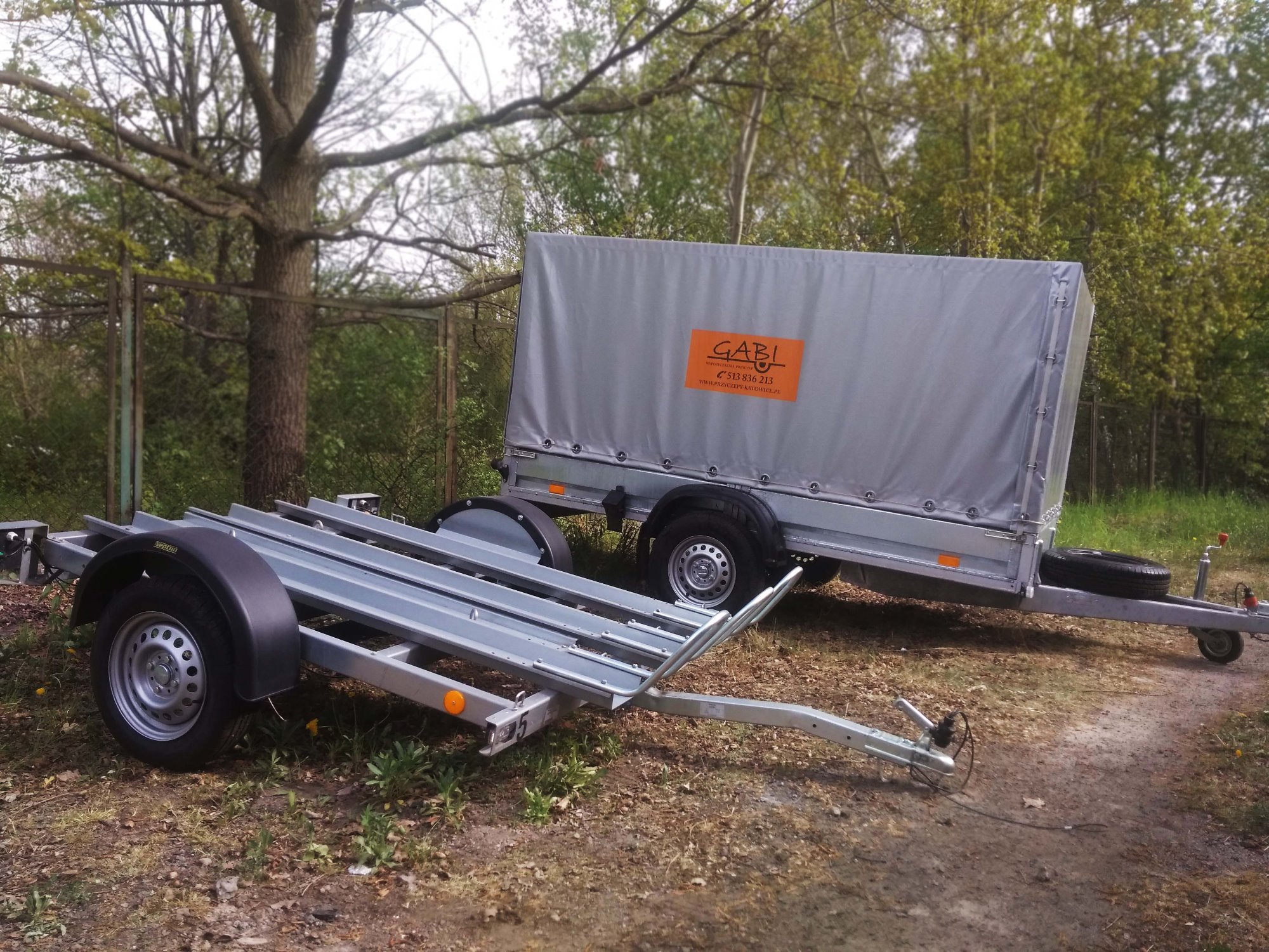 Dwie przyczepy samochodowe na trawiastym terenie; jedna z plandeką z pomarańczowym logo firmy, druga bez plandeki, z konstrukcją do transportu motocykli.