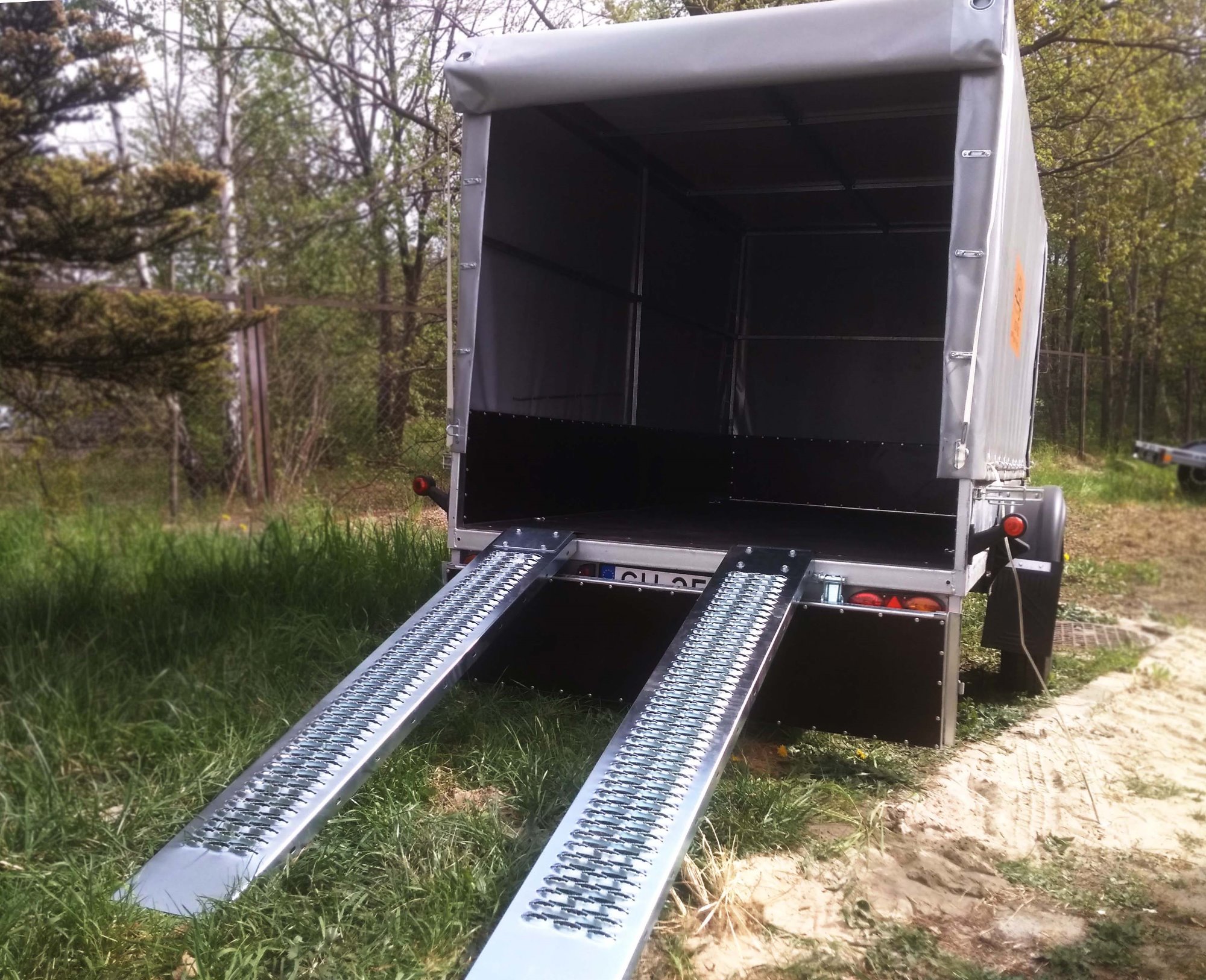 Otwarta przyczepa z najazdami, gotowa do załadunku, widok z tyłu na tle zieleni.
