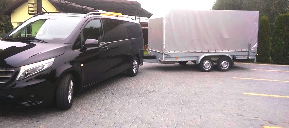Czarny van Mercedes z przyczepą krytą plandeką na parkingu z kostki brukowej.