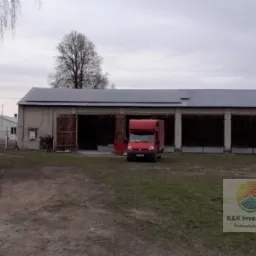 Duży budynek gospodarczy z zamontowanymi panelami słonecznymi na dachu, obok zaparkowany czerwony samochód dostawczy z otwartymi drzwiami, widoczny znak firmowy w prawym dolnym rogu.