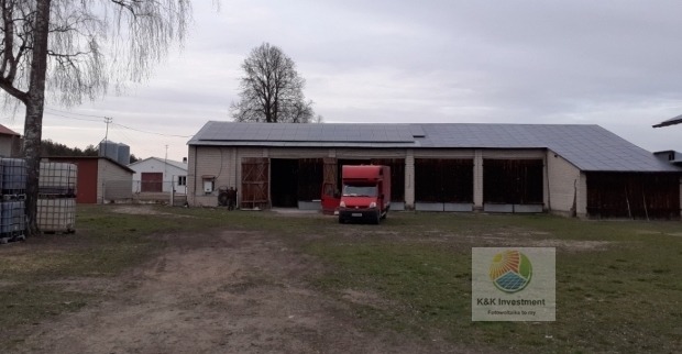 Duży budynek gospodarczy z zamontowanymi panelami słonecznymi na dachu, obok zaparkowany czerwony samochód dostawczy z otwartymi drzwiami, widoczny znak firmowy w prawym dolnym rogu.