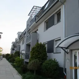 Szeregowy budynek mieszkalny z szarą elewacją, balkonami z metalowymi balustradami i daszkami nad balkonami oraz wejściami, widok z chodnika z zaparkowanymi samochodami i zielenią.