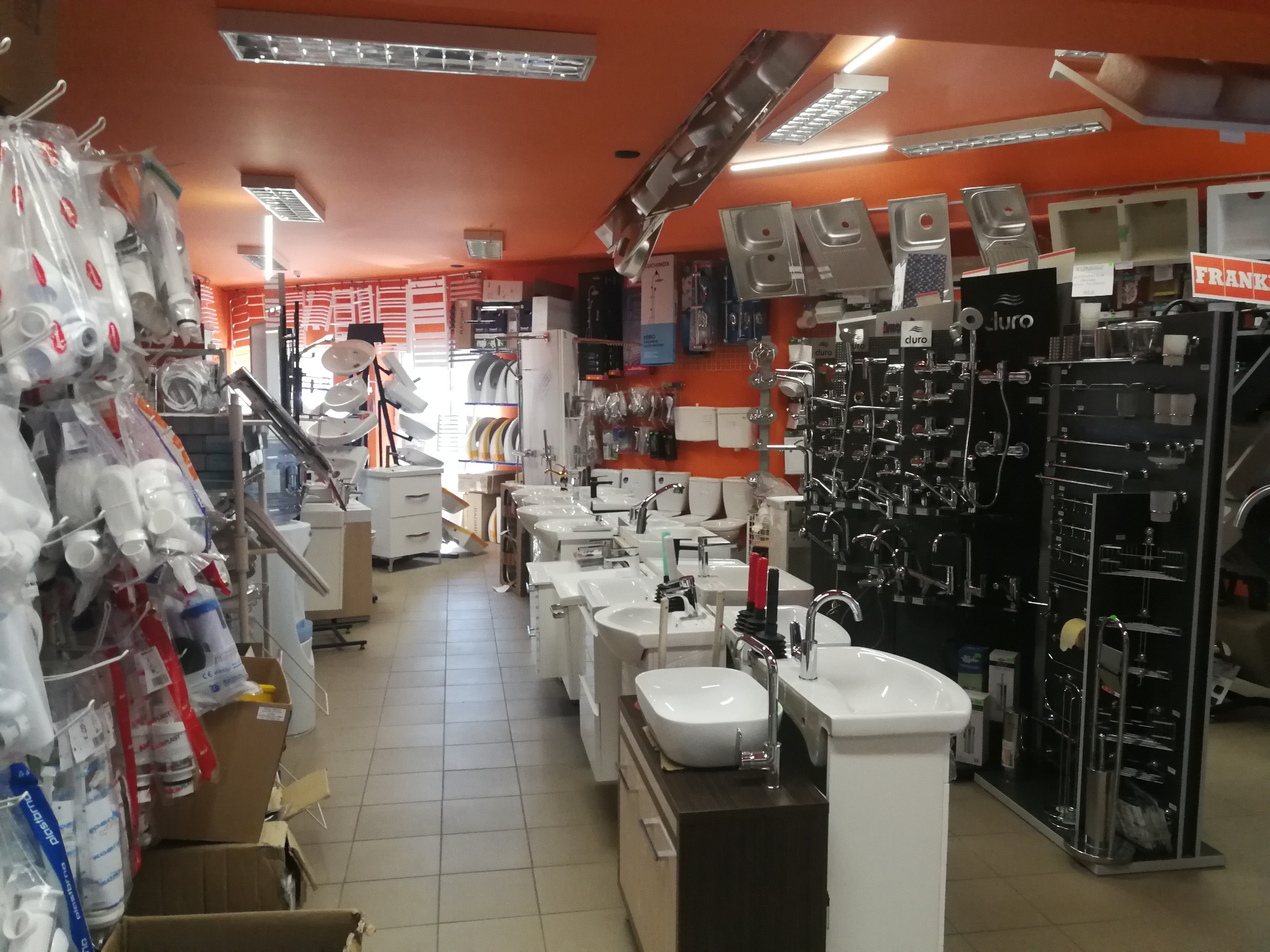 Wnętrze sklepu z armaturą łazienkową, prezentujące umywalki nablatowe na szafkach, baterie, zlewozmywaki i rury PCV w foliowych opakowaniach, oświetlone jarzeniówkami.