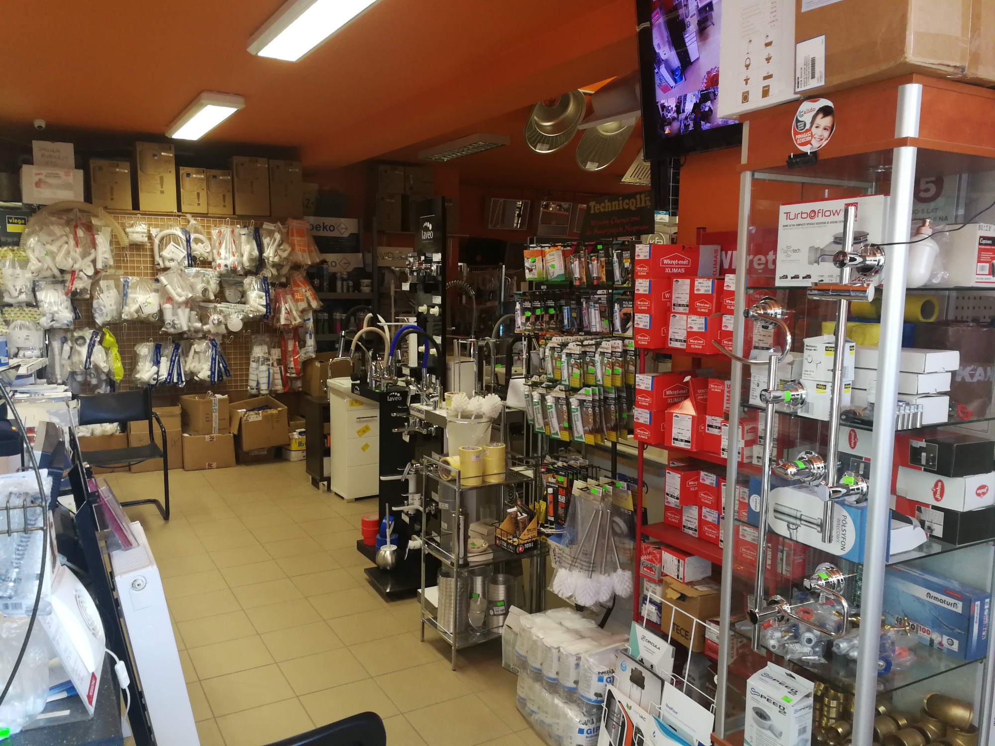 Wnętrze sklepu z armaturą i akcesoriami hydraulicznymi, widoczne półki z bateriami, rurami, złączkami, środkami chemicznymi i narzędziami, oświetlenie sufitowe i pomarańczowe ściany.