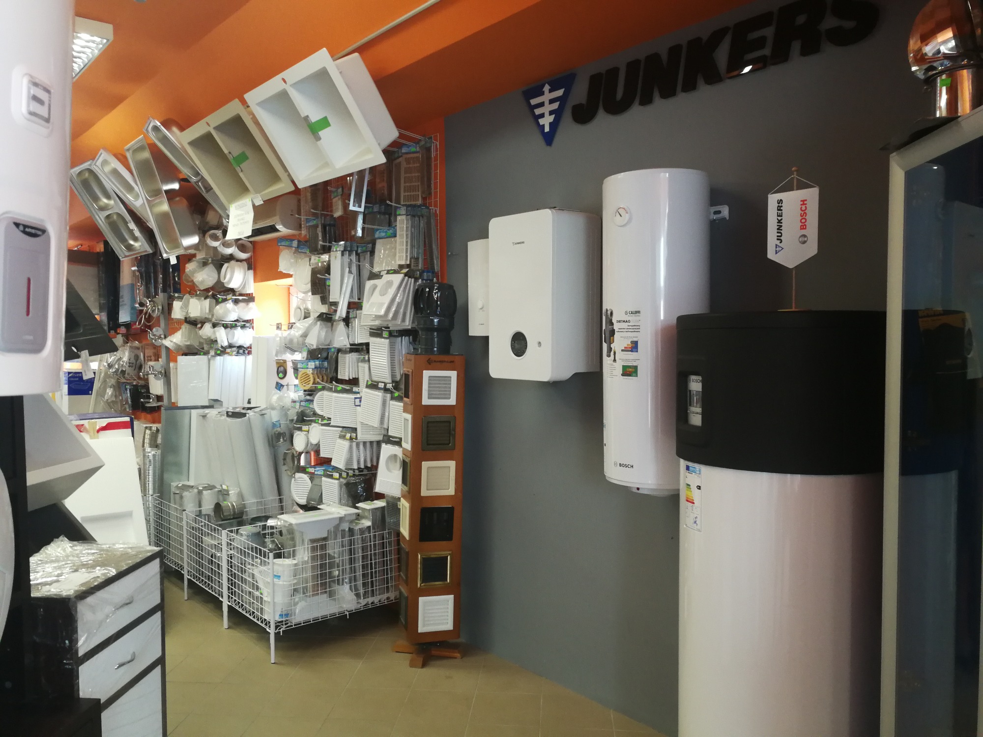 Wystawa w sklepie instalacyjnym: białe umywalki, kratki wentylacyjne na obrotowym stojaku, bojlery wiszące i stojące marek Junkers i Bosch, firmowa flaga Junkers-Bosch. Pomarańczowy sufit.