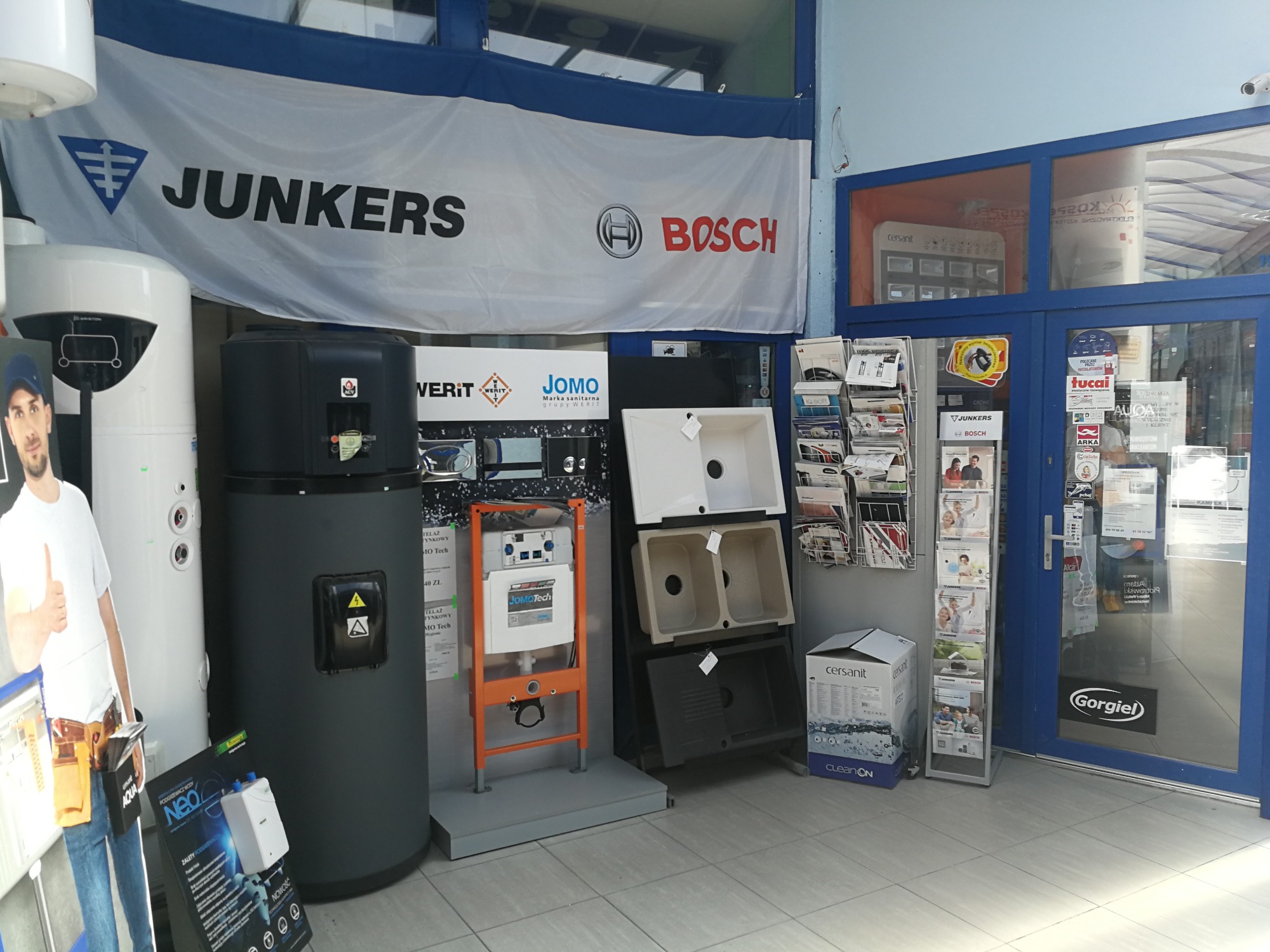 Wnętrze sklepu hydraulicznego z ekspozycją urządzeń sanitarnych, w tym umywalek, systemów podtynkowych i podgrzewaczy wody; baner reklamowy Junkers i Bosch w tle.