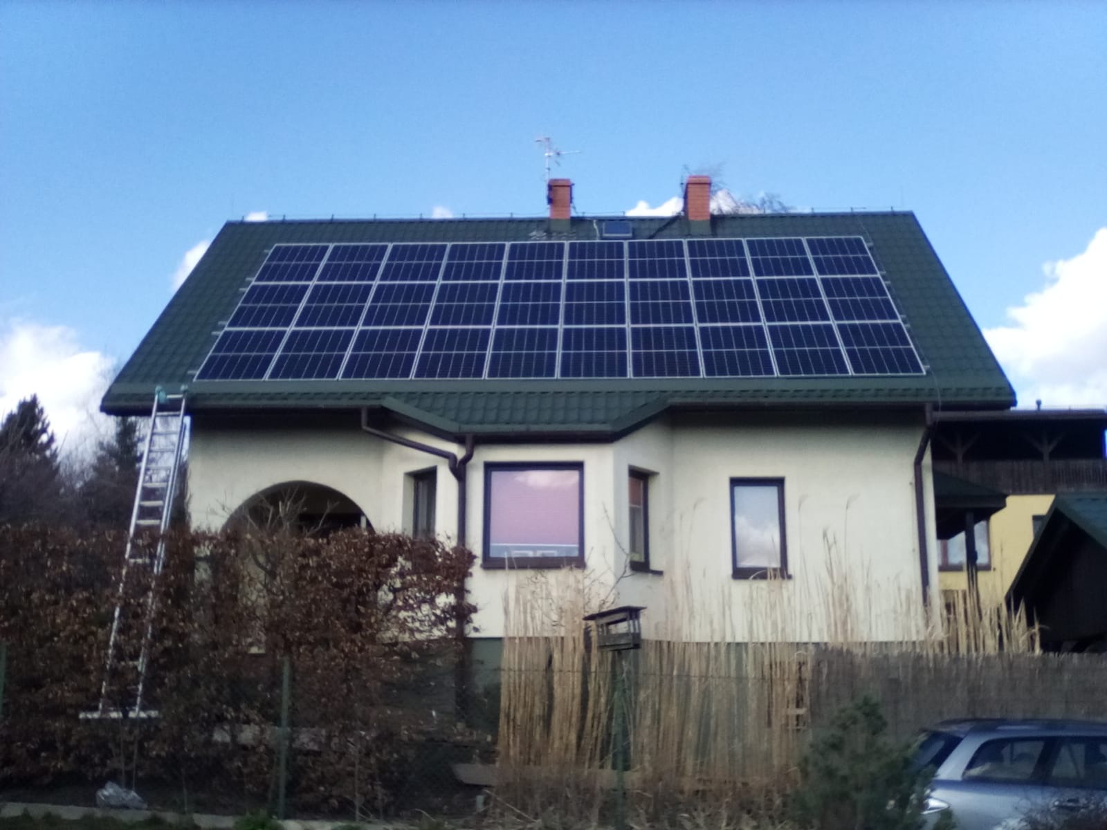 Dom jednorodzinny z panelami słonecznymi na dachu z zielonej blachy, drabina oparta o rynnę, widok z poziomu gruntu zza ogrodzenia.