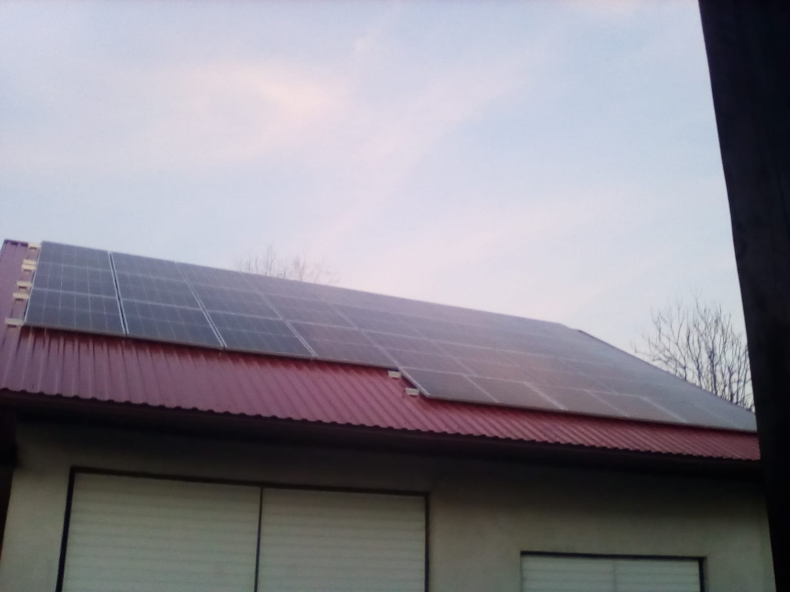 Panele słoneczne na czerwonym dachu budynku z szarą elewacją i zamkniętymi roletami antywłamaniowymi, widoczne drzewa bez liści w tle.