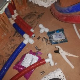 Instalatorstwo Sanitarne i Gazowe - Fragment instalacji wodnej w trakcie montażu w Warszawie: czerwone i niebieskie rury, złączki, narzędzia do cięcia rur oraz opakowania elementów instalacyjnych na surowym podłożu.