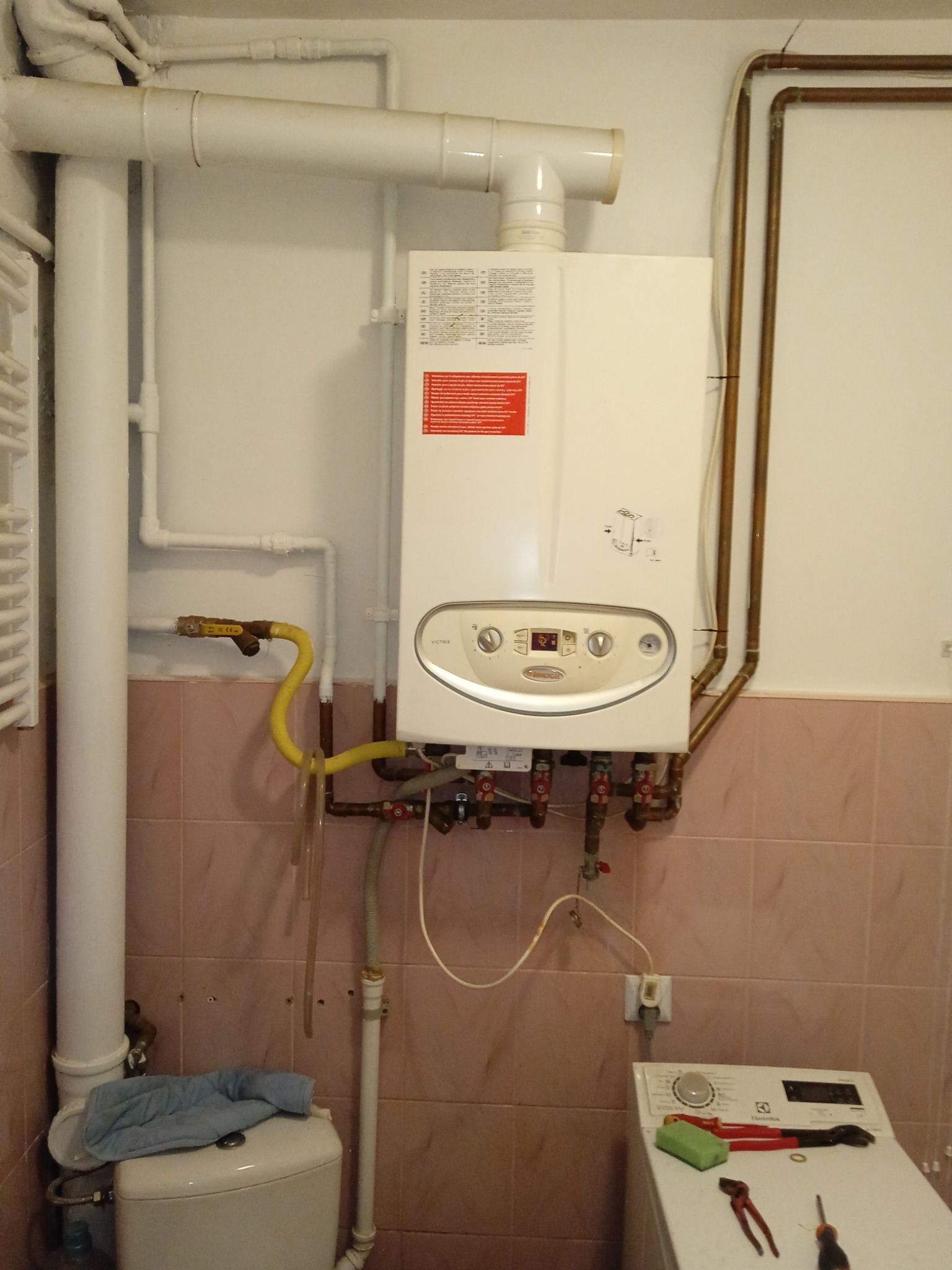 Biały piec gazowy zamontowany na ścianie wyłożonej różowymi płytkami, widoczne rury gazowe, zawory, przewody oraz pralka z narzędziami obok. Niechlujne otoczenie.