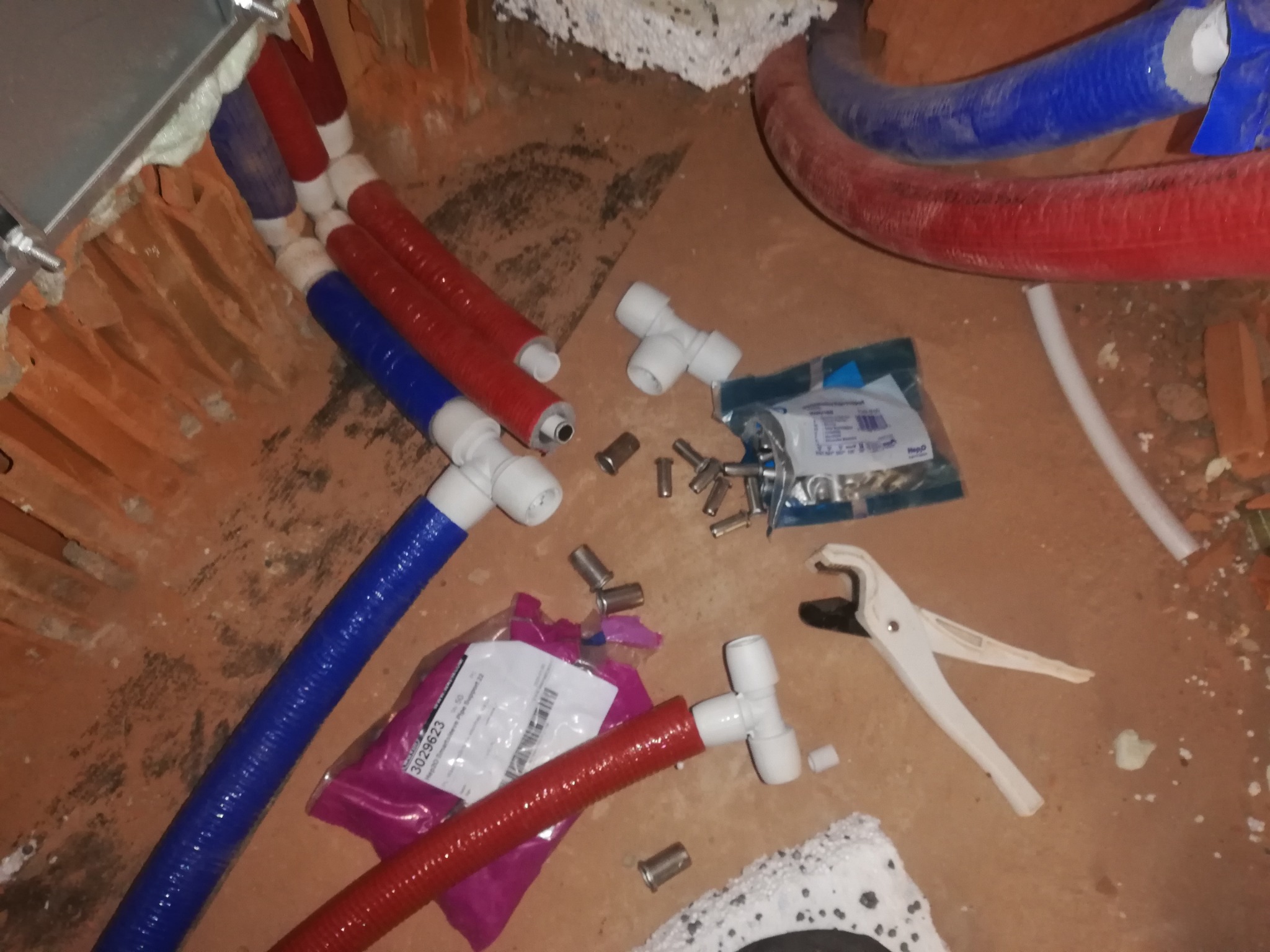 Fragment instalacji wodnej w trakcie montażu w Warszawie: czerwone i niebieskie rury, złączki, narzędzia do cięcia rur oraz opakowania elementów instalacyjnych na surowym podłożu.