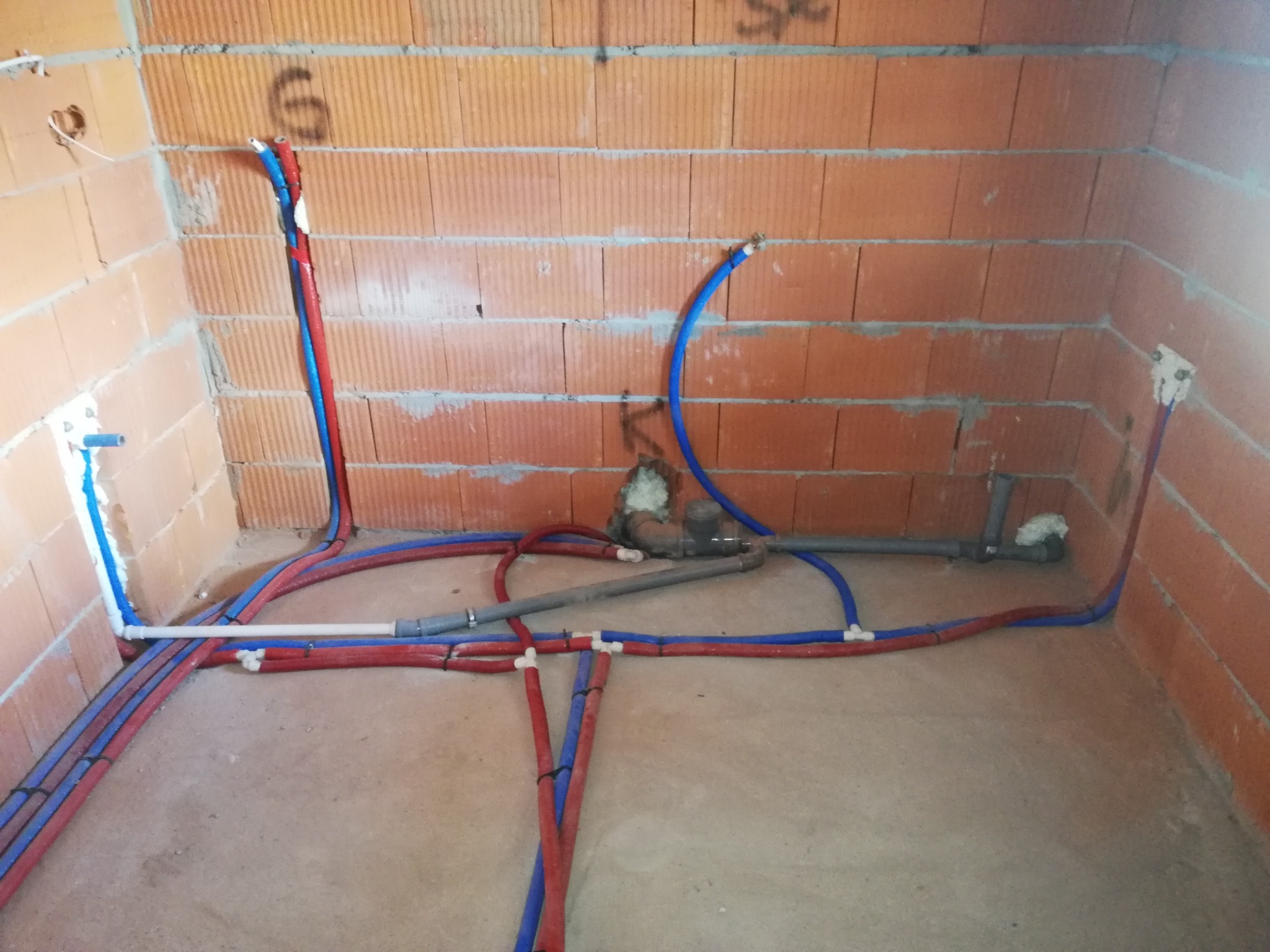 Surowe wnętrze z ceglanymi ścianami w trakcie instalacji rur wodnych, czerwone i niebieskie, z widocznymi złączkami i fragmentami szarej kanalizacji.