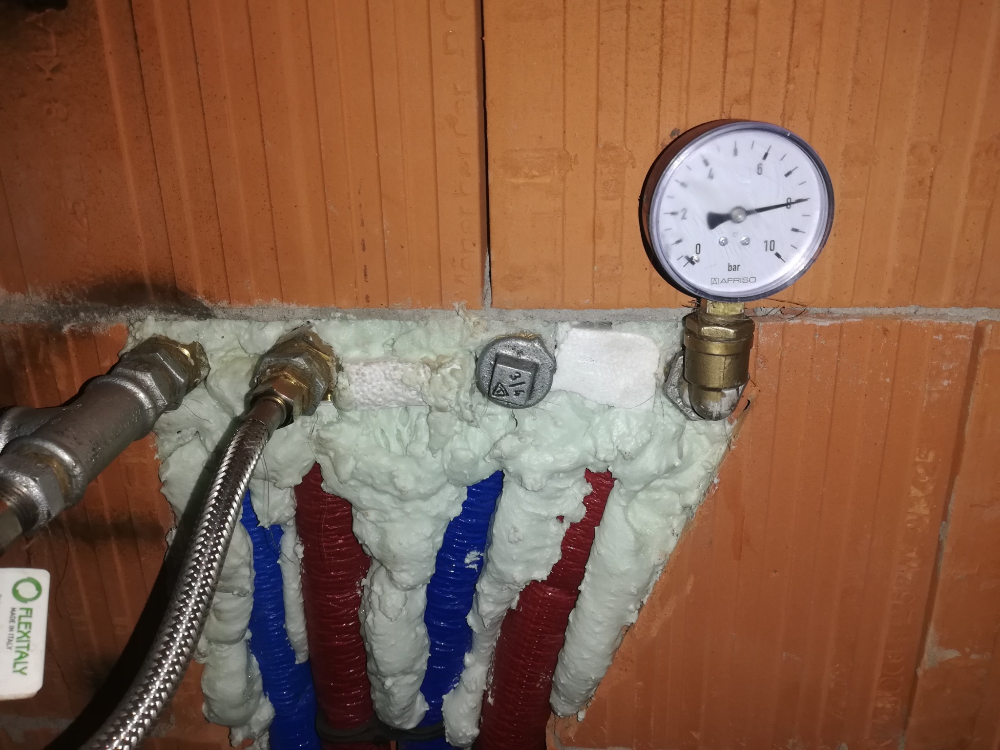 Podłączenie rur grzewczych w ścianie z cegły, zabezpieczone pianką montażową, z widocznym manometrem AFRISO i fragmentem wężyka FEXTALY.