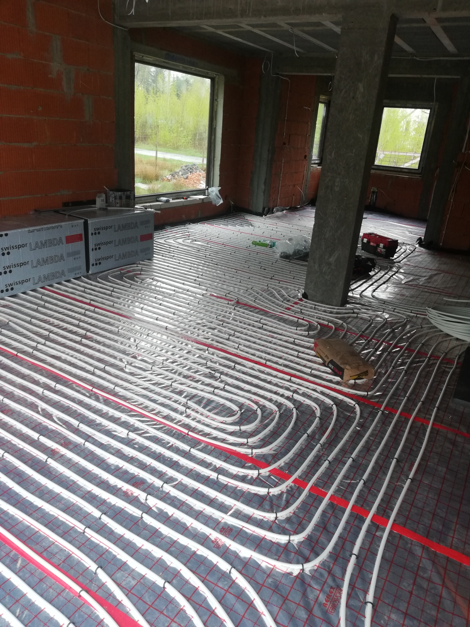Rozłożona instalacja ogrzewania podłogowego w surowym stanie budynku, widoczne rury grzewcze na folii z siatką, z oknem w tle i materiałami budowlanymi obok.