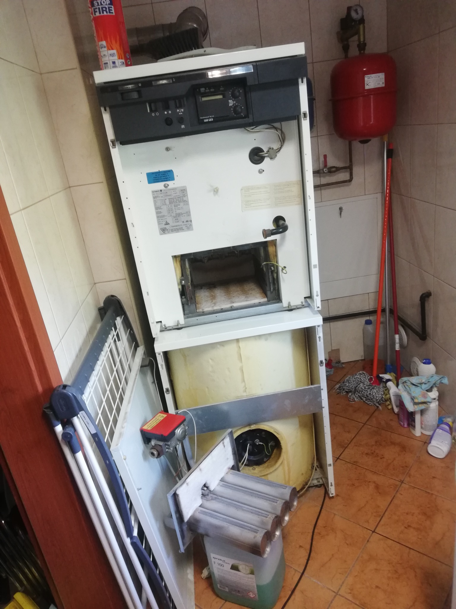 Otwarty piec gazowy w trakcie demontażu lub naprawy, z widocznymi elementami wewnętrznymi i zewnętrznymi, butla gaśnicza na górze, bałagan na podłodze.