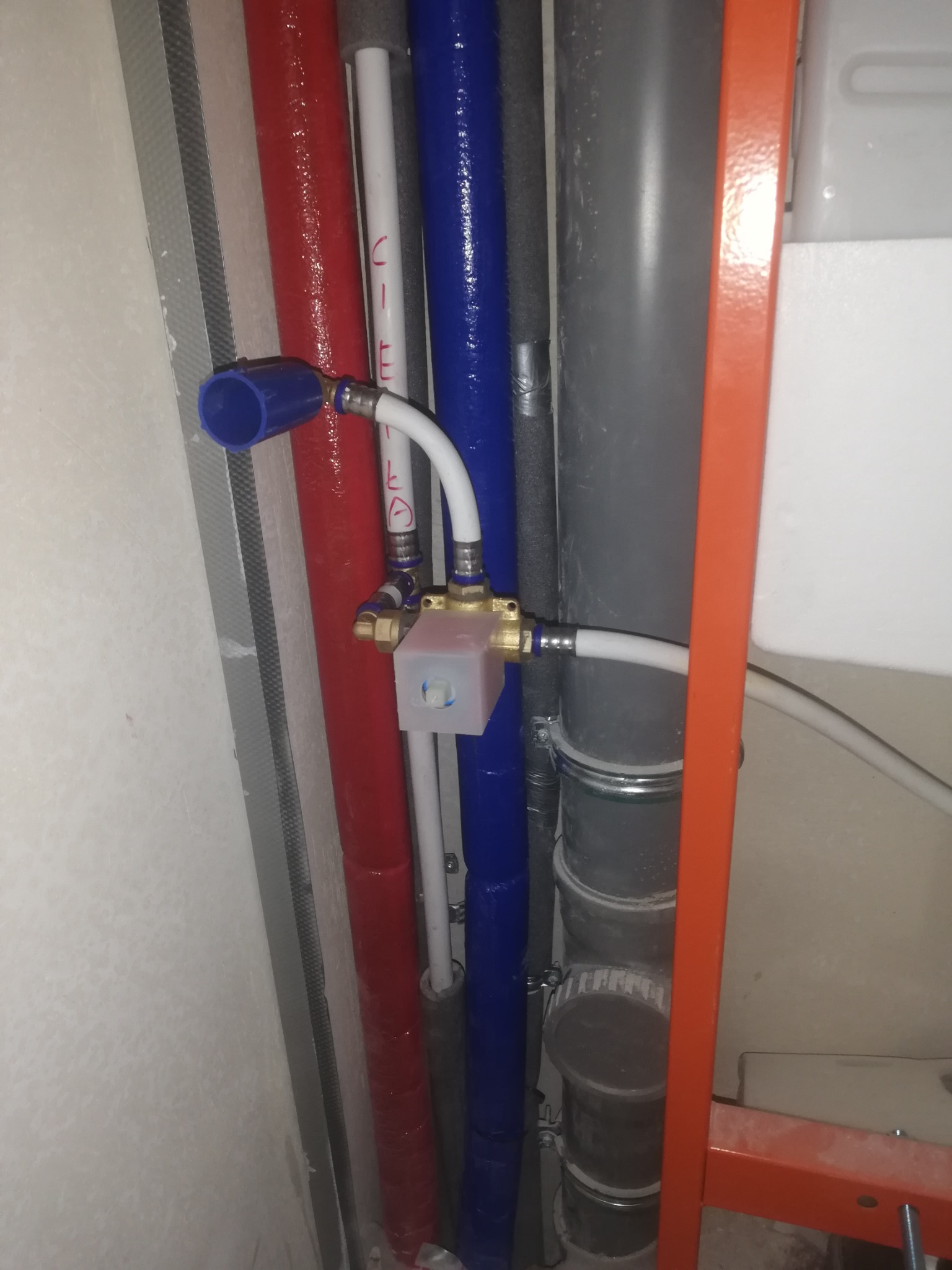 Różnokolorowe rury instalacyjne (czerwona, biała, niebieska, szara) z zaworem termostatycznym w surowym stanie, widoczne elementy mocujące i izolacja.