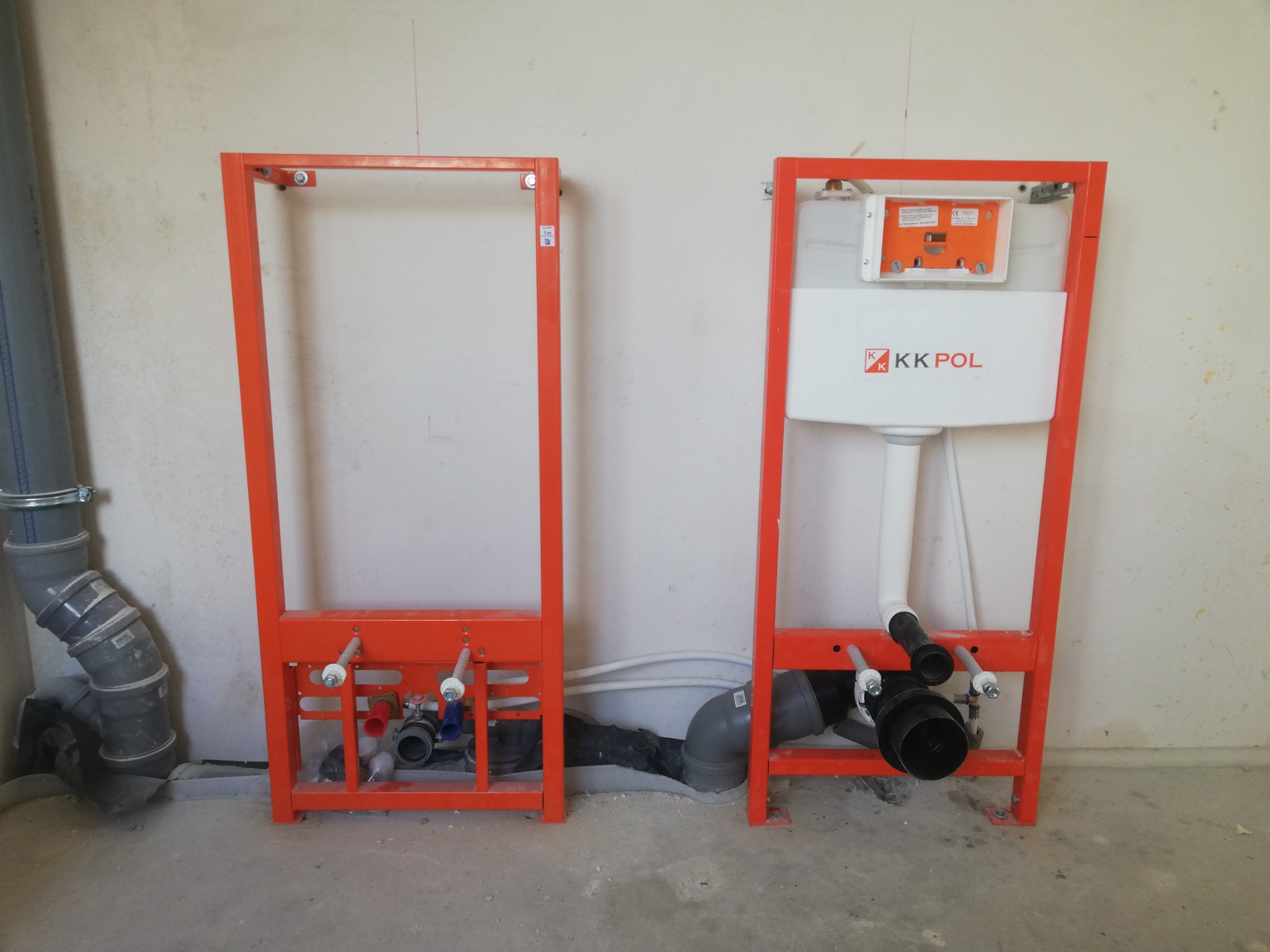 Dwa pomarańczowe stelaże podtynkowe do WC, jeden z zamontowaną spłuczką KK-POL, widoczne rury kanalizacyjne i przyłącza wodne, surowa ściana w tle.
