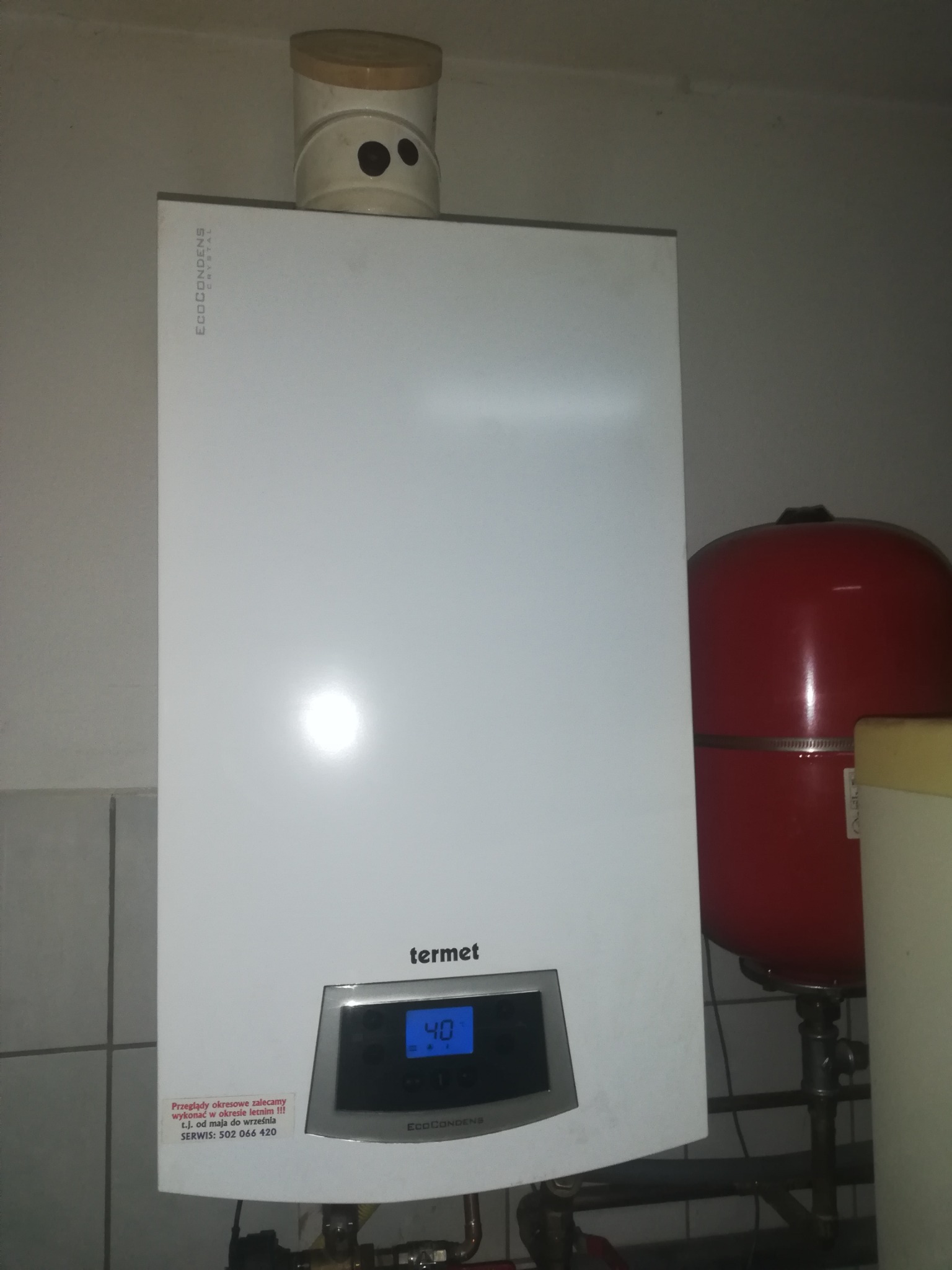 Biały, wiszący piec gazowy Termet EcoCondens Crystal z wyświetlaczem wskazującym 40 stopni, czerwony zbiornik wyrównawczy obok, widoczna naklejka z numerem serwisu, w tle kafelki.