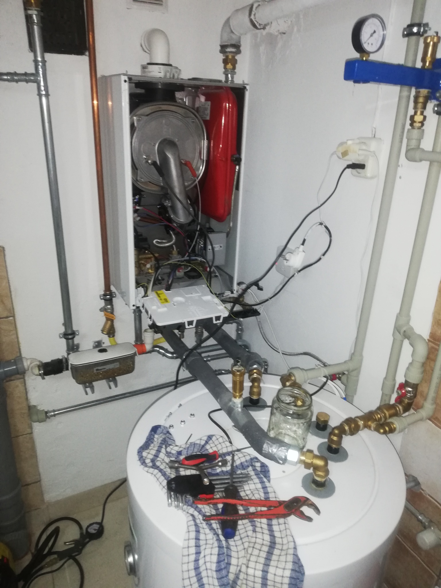 Otwarty piec gazowy z widocznymi elementami wewnętrznymi, podłączony do instalacji, z narzędziami i słoikiem na zbiorniku, w trakcie prac instalacyjnych.