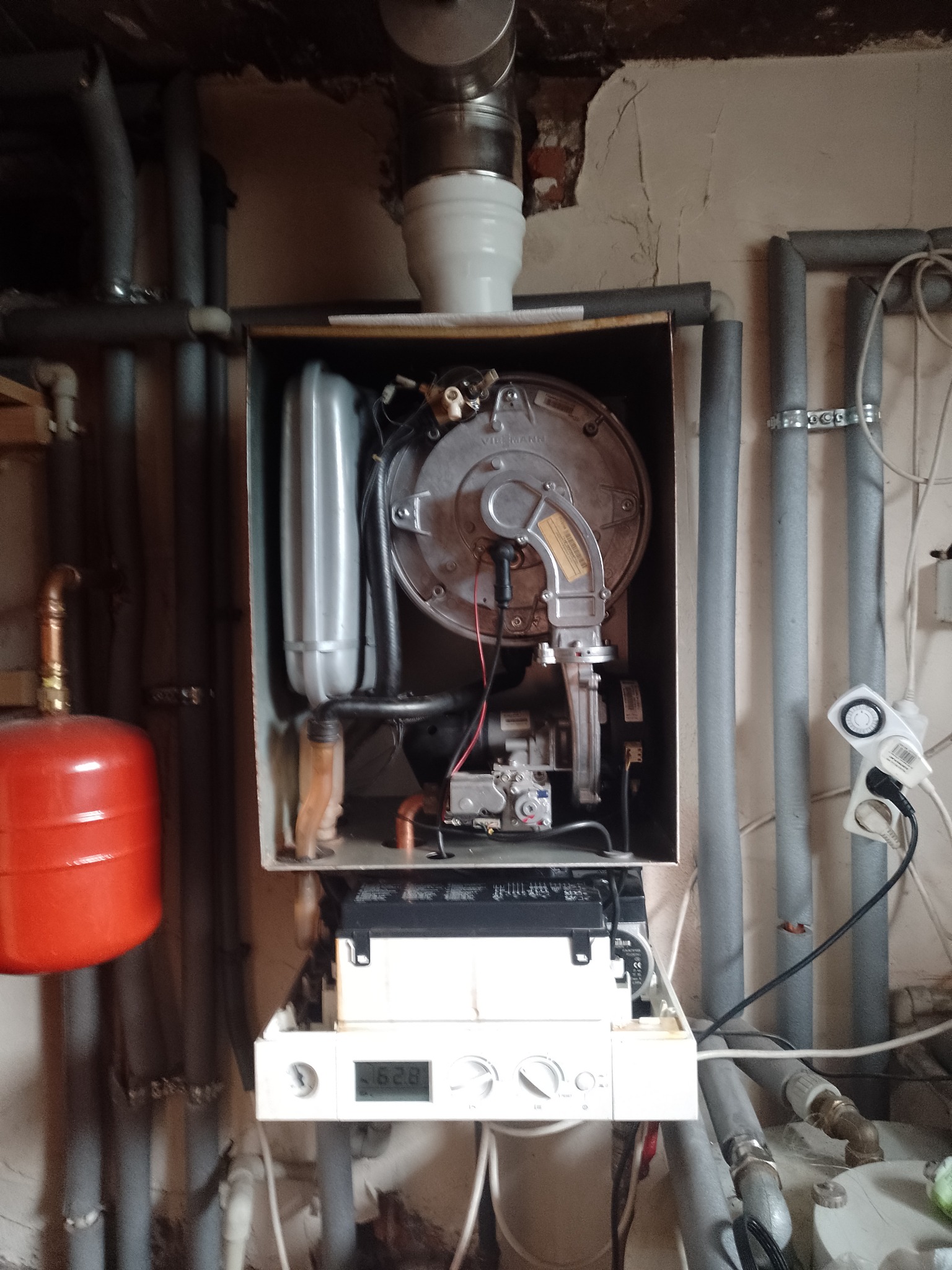 Otwarty piec gazowy Viessmann z widocznymi elementami wewnętrznymi, obok rury centralnego ogrzewania i czerwony zbiornik wyrównawczy, na tle ściany z widocznymi śladami użytkowania.