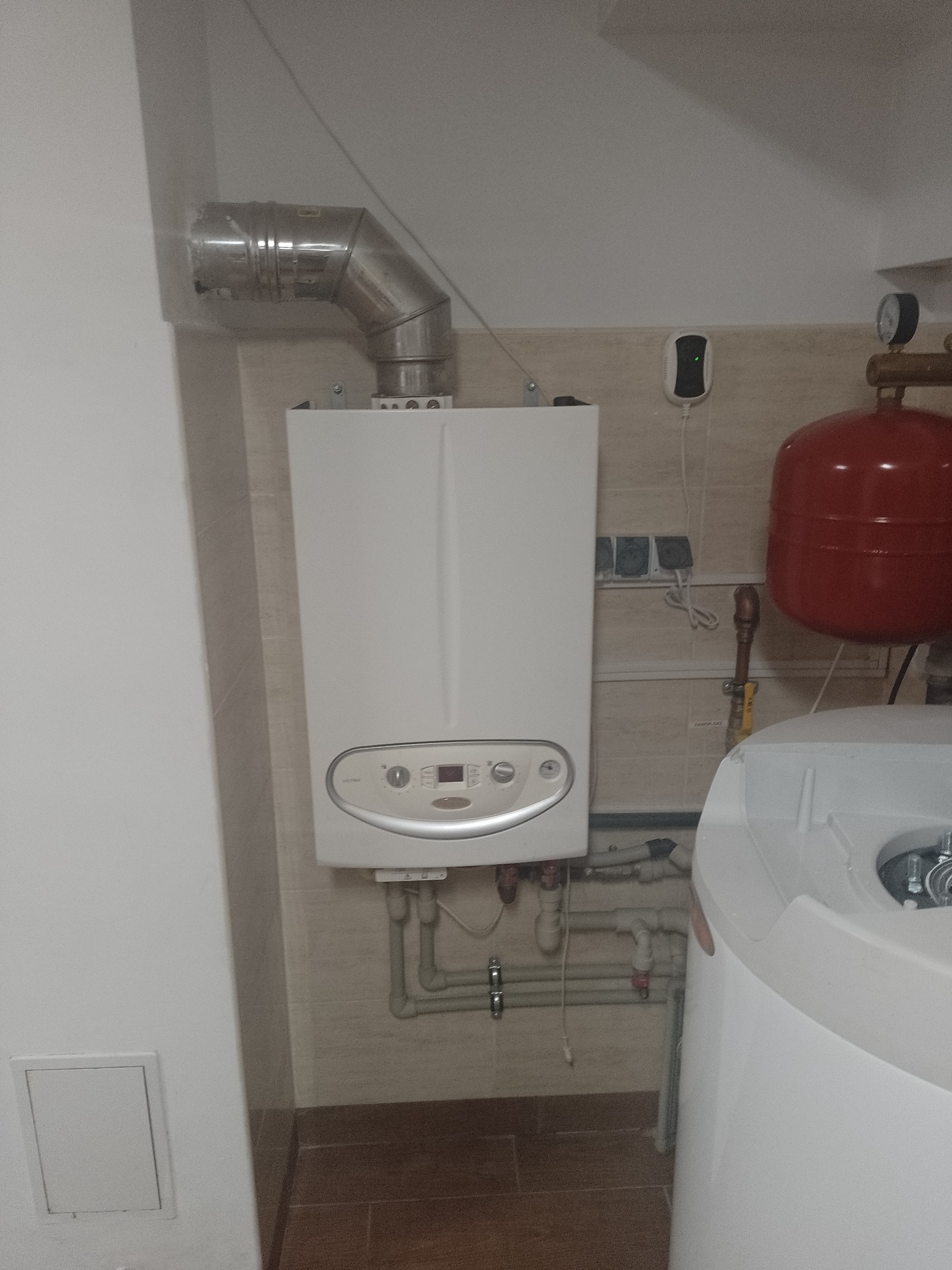 Biały piec gazowy z podłączonym kominem i instalacją hydrauliczną, widoczny zbiornik wyrównawczy, zamontowany na tle beżowych płytek.