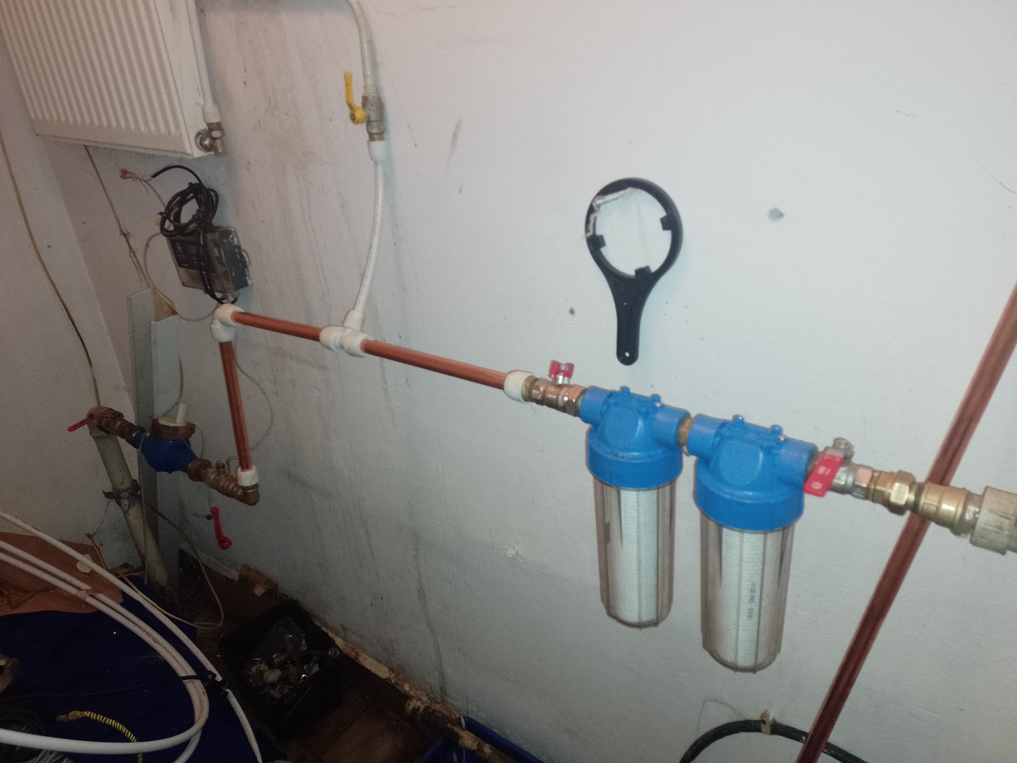 Dwa filtry wody z niebieskimi obudowami zamontowane na miedzianych rurach, widoczne zawory odcinające i klucz do filtrów na ścianie, w tle grzejnik.