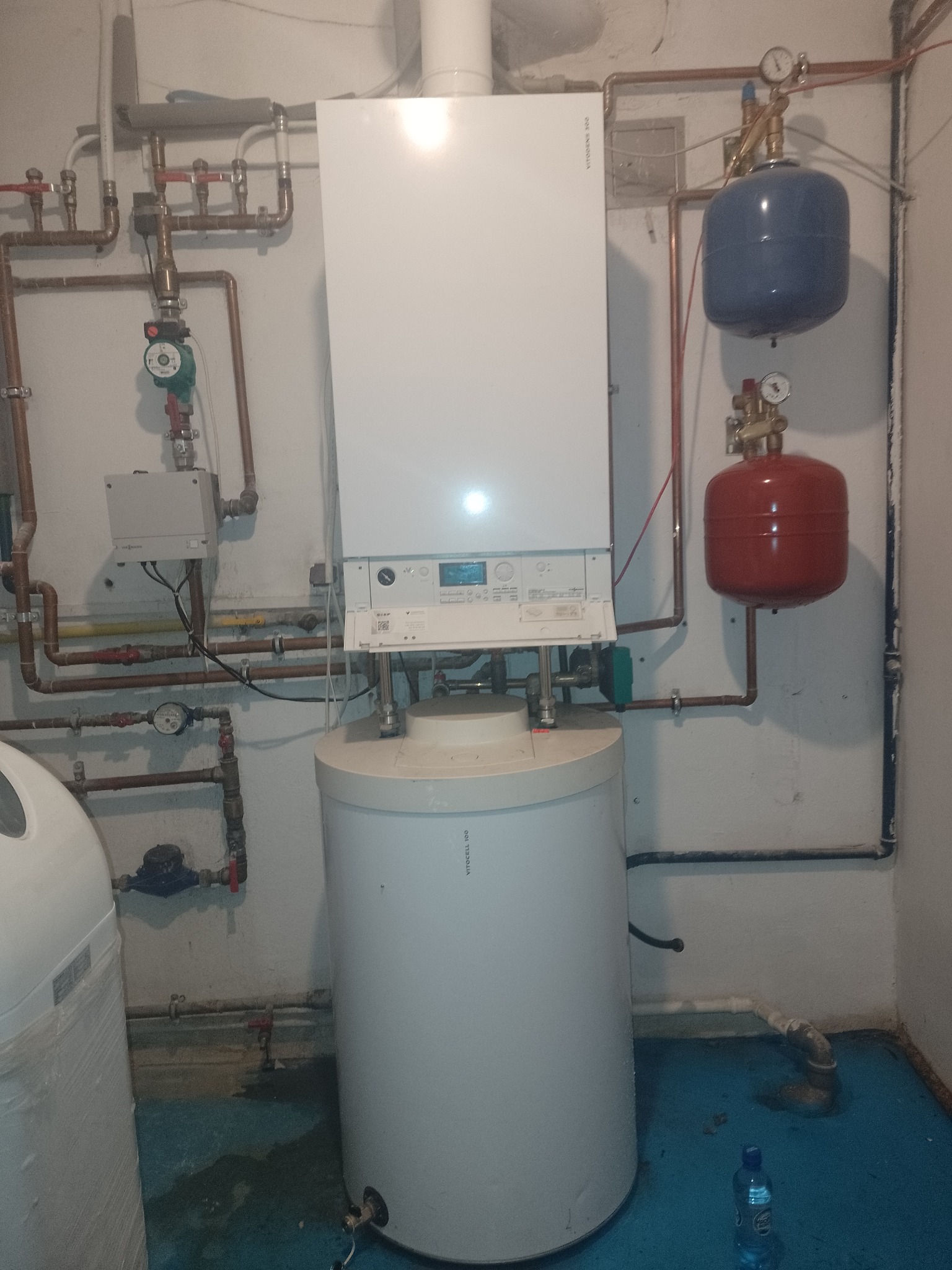 Kompaktowa kotłownia z białym piecem gazowym Viessmann, zbiornikiem ciepłej wody, niebieskim i czerwonym naczyniem przeponowym, oraz miedzianą instalacją grzewczą w pomieszczeniu z niebieską podłogą.