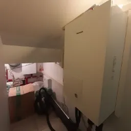 Biała pompa ciepła Panasonic zamontowana w pomieszczeniu technicznym, widoczne rury i zawory, w tle kartonowe pudło i odkurzacz.