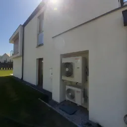 Dwie jednostki zewnętrzne klimatyzacji Toshiba Inverter zamontowane na białej ścianie nowoczesnego domu, widok z boku na tle zielonego trawnika i błękitnego nieba.