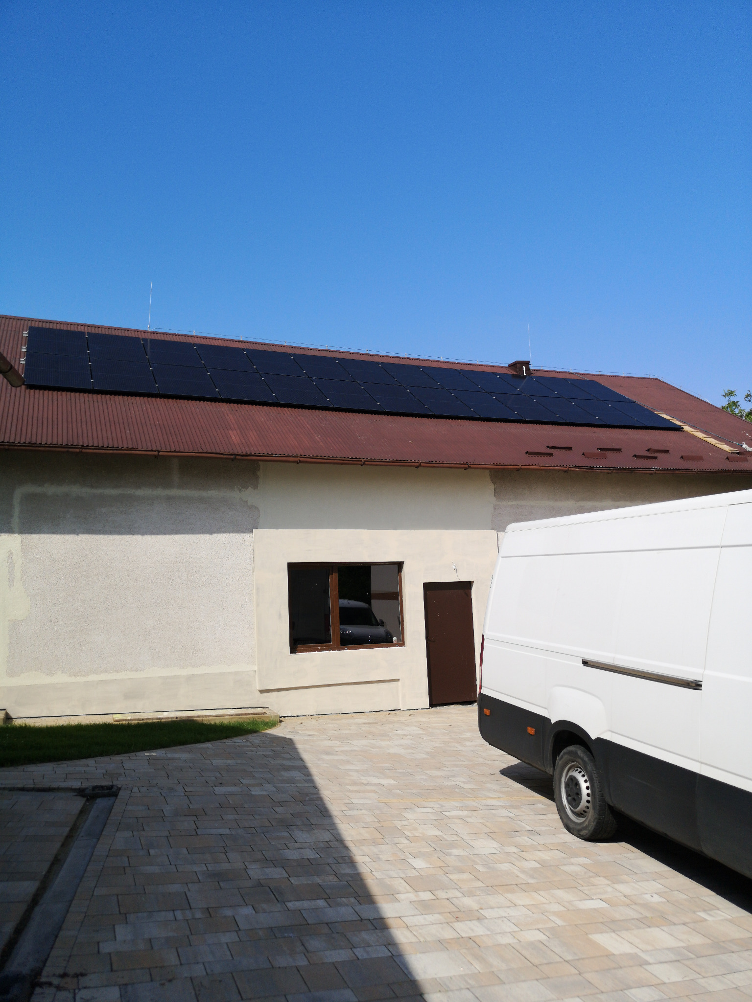 Panele słoneczne na brązowym dachu budynku z kremową elewacją, obok zaparkowany biały bus dostawczy.
