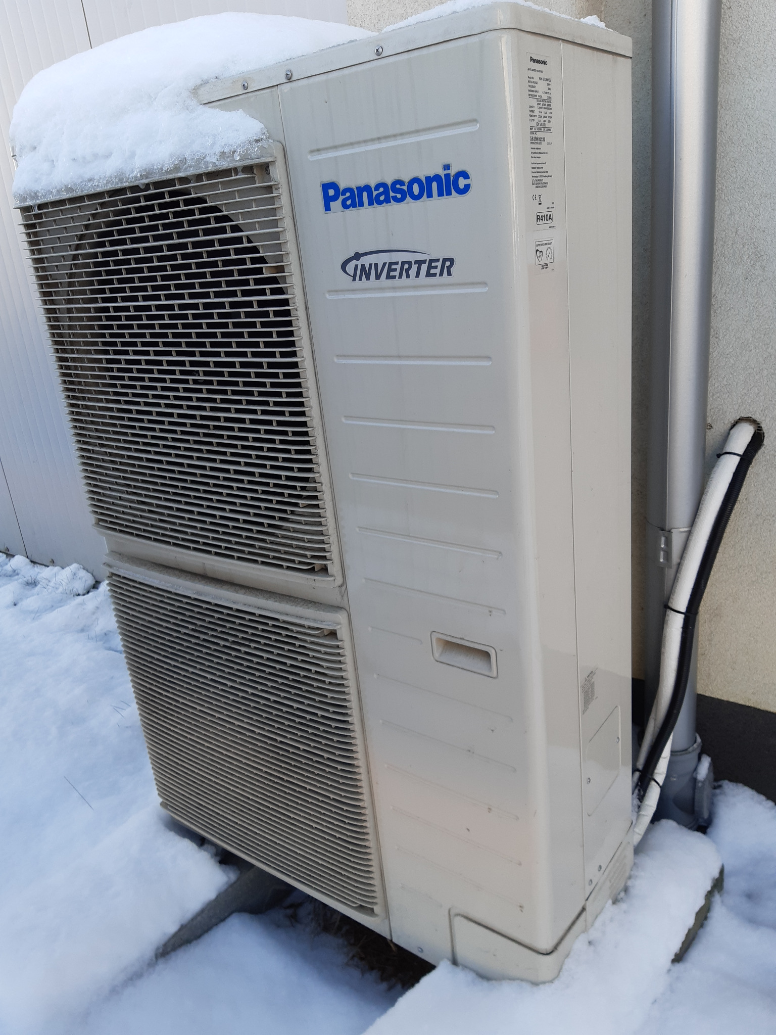 Zewnętrzna jednostka klimatyzacji Panasonic Inverter, częściowo przysypana śniegiem, z widocznymi rurami instalacyjnymi i fragmentem ściany budynku.