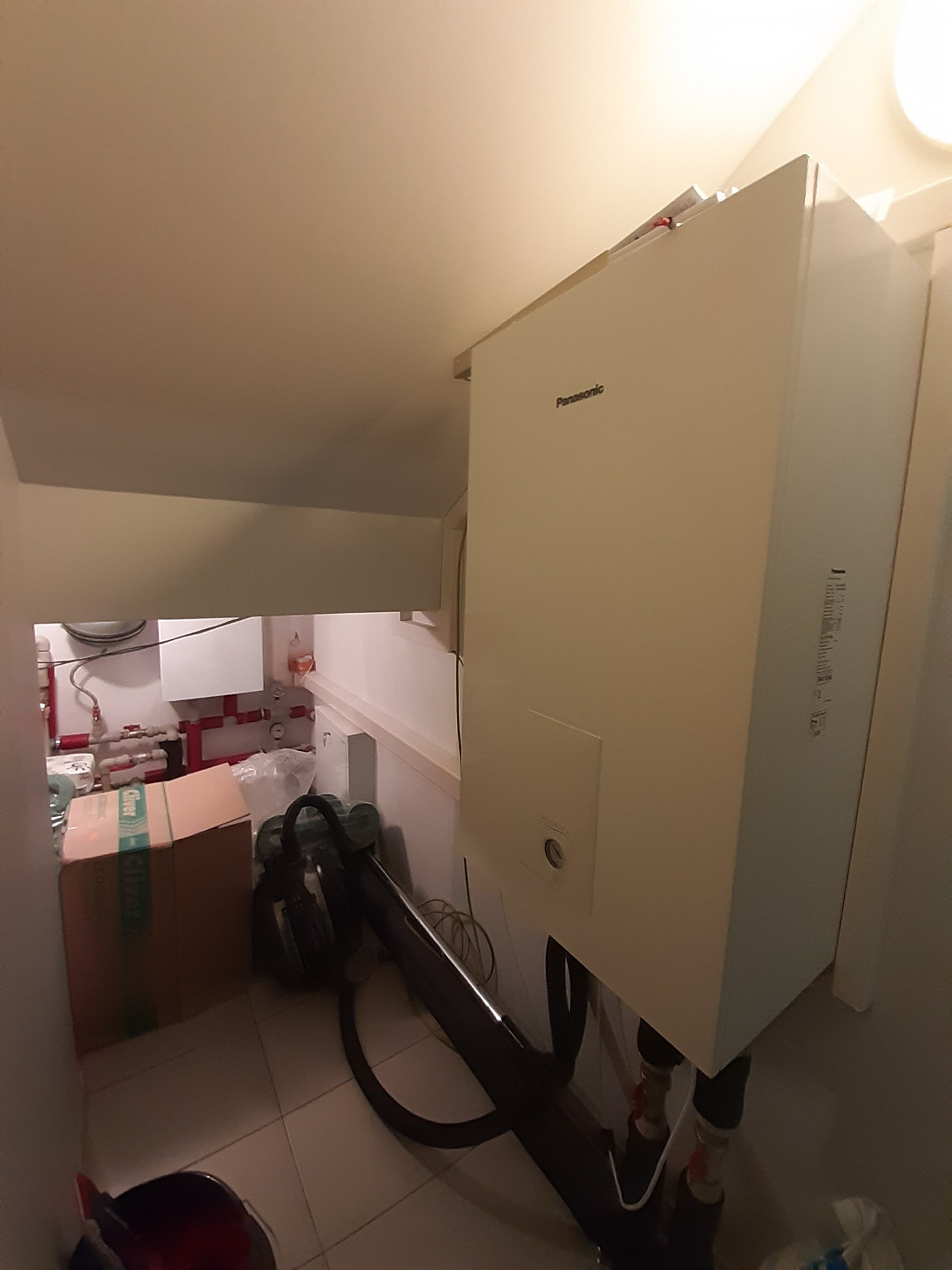 Biała pompa ciepła Panasonic zamontowana w pomieszczeniu technicznym, widoczne rury i zawory, w tle kartonowe pudło i odkurzacz.