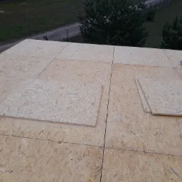 Montaż płyt OSB na dachu budynku, widoczna piła elektryczna i poziomica, w tle drzewa i ogrodzenie.