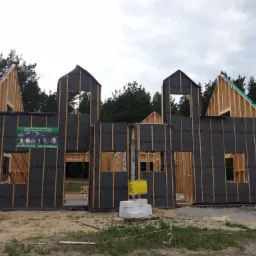 Surowy szkielet drewnianego domu w trakcie budowy, zabezpieczony czarną membraną, widoczne belki konstrukcyjne i fragmenty dachu w tle las.